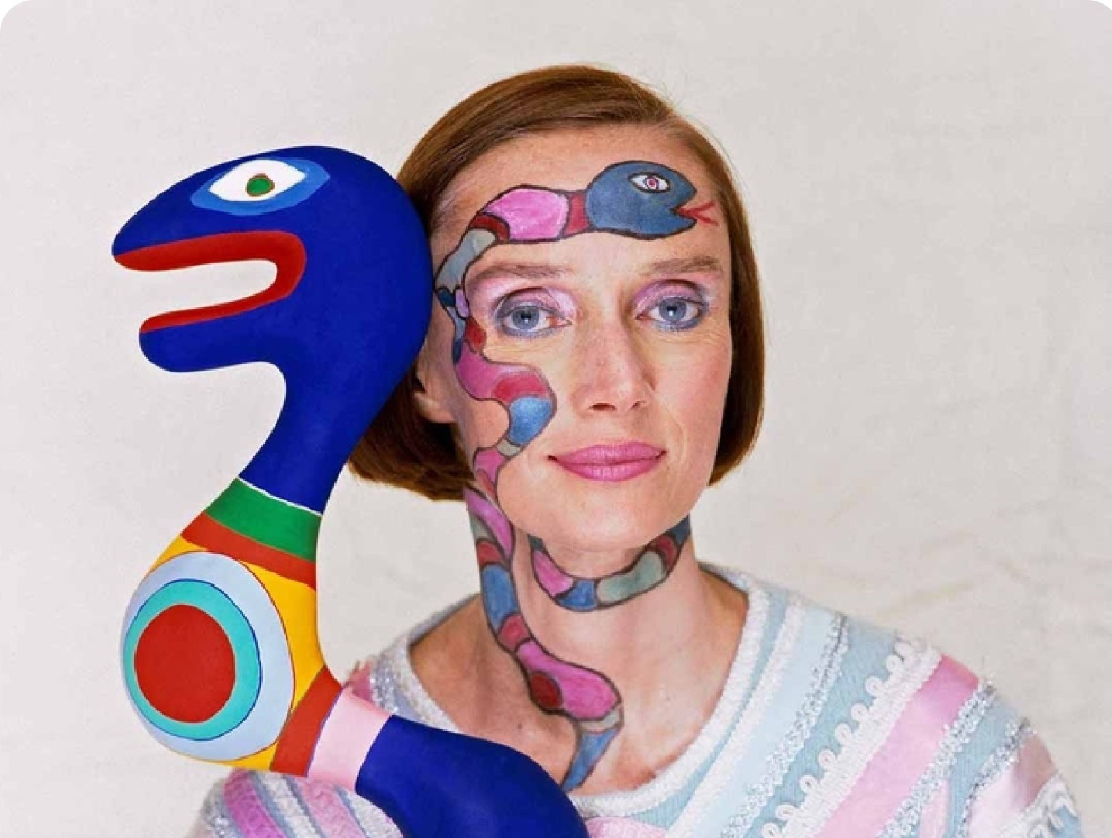 Artista plástica Niki de Saint Phalle está de frente e olha para a câmera segurando a seu lado escultura azul de criatura parecida com animal