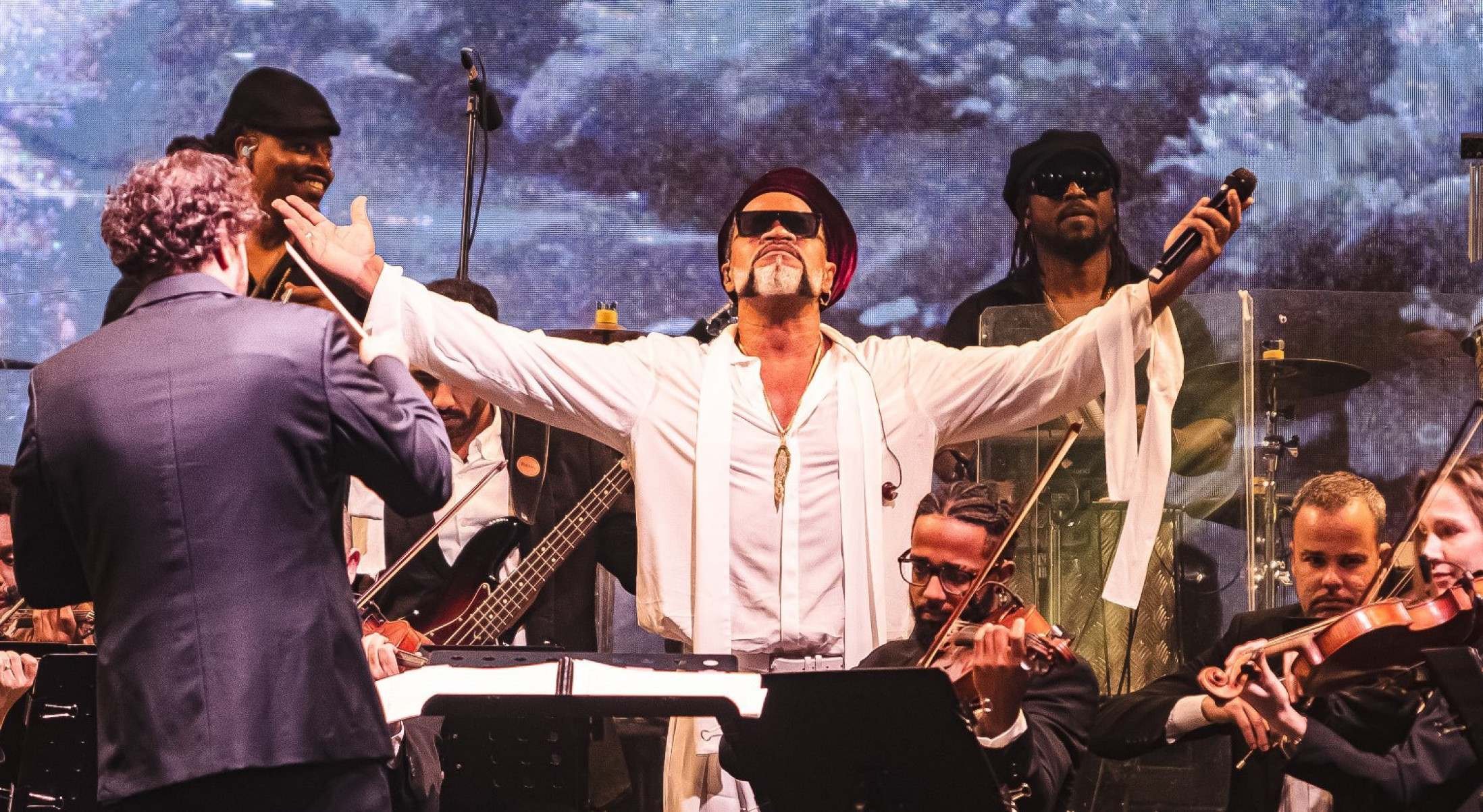 Cantor Carlinhos Brown está com os dois braços abertos e atrás dele estão músicos e maestros da Orquestra Ouro Preto