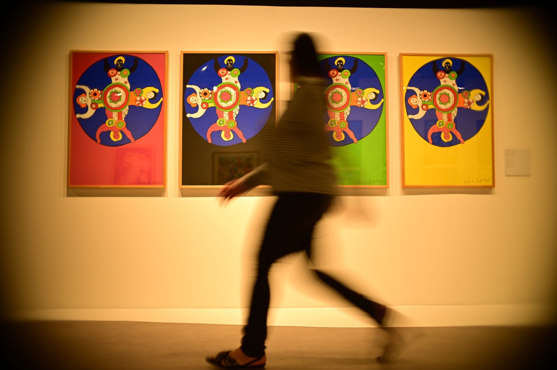 Pessoa passa em frente à série de quadros muito coloridos de Niki Saint Phalle expostos em Belo Horizonte