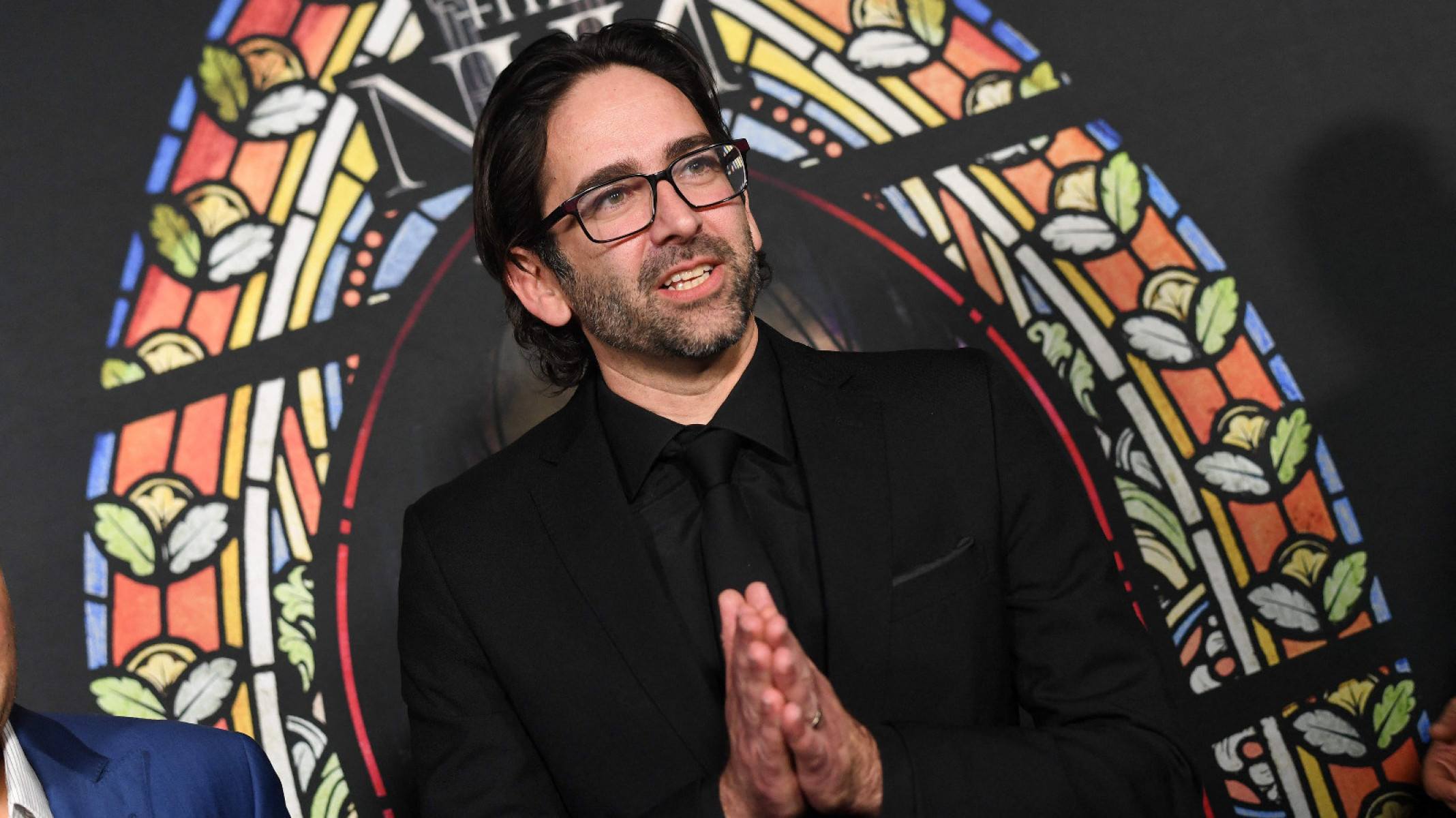 Diretor de cinema Michael Chaves sorri, atrás dele há um vitral colorido