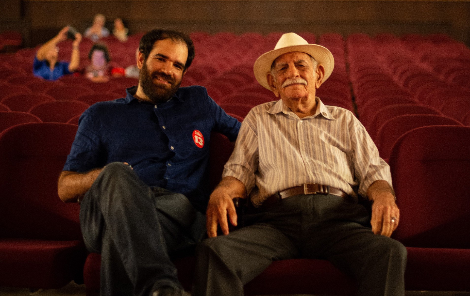 O cineasta Leonardo Lacca com o avô, Seu Cavalcanti, protagonista do filme lançado pelo neto.Os dois sorriem para a câmera e estão sentados em uma sala de cinema