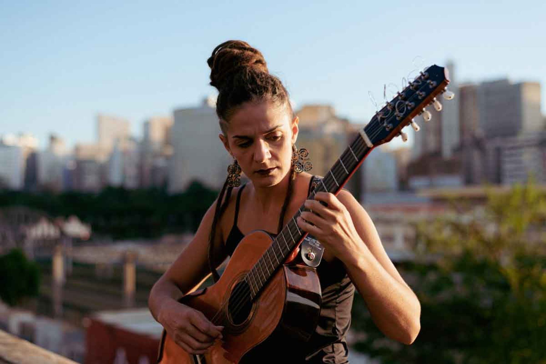 A cantora e compositora Letícia Leal está tocando um violão e ao fundo da foto há prédios e fotos desfocados. Ela usa brincos grandes em formato de folha, um rabo de cavalo com as tranças afro caindo do lado esquerdo do ombro e camiseta marrom. 