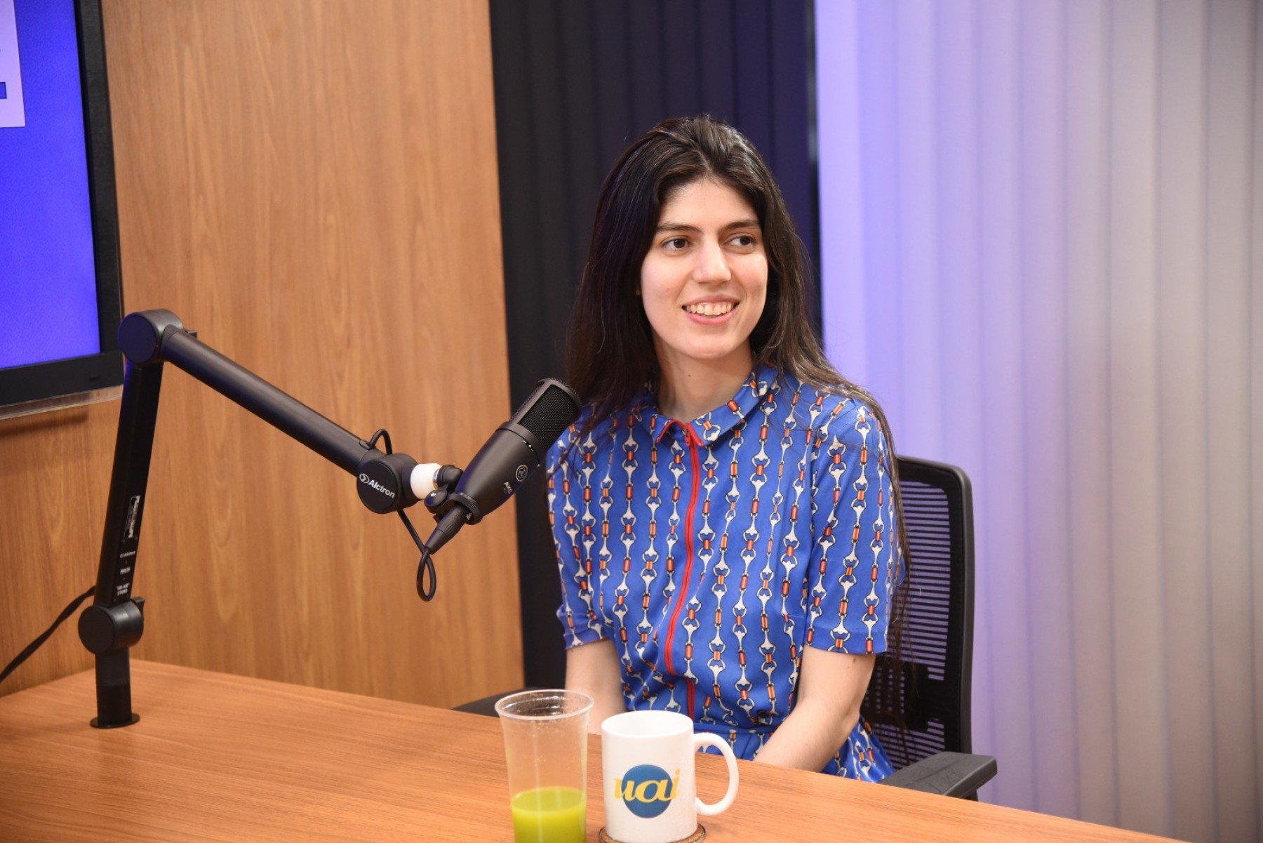 Moça de cabelos pretos, blusa azul  sentada à meda de um estúdio de podcast