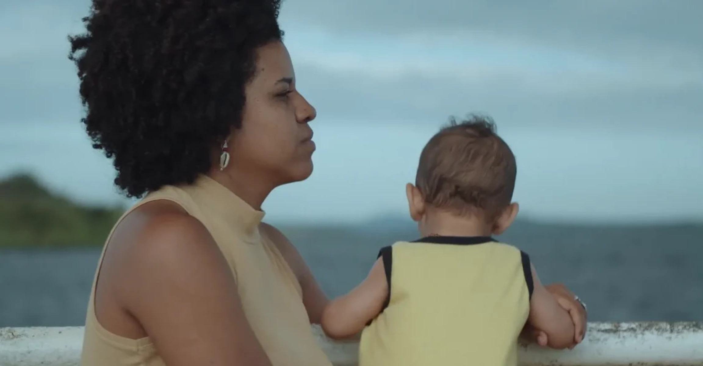 Mulher segura criança e tem ao fundo um rio, em cena do filme Cais