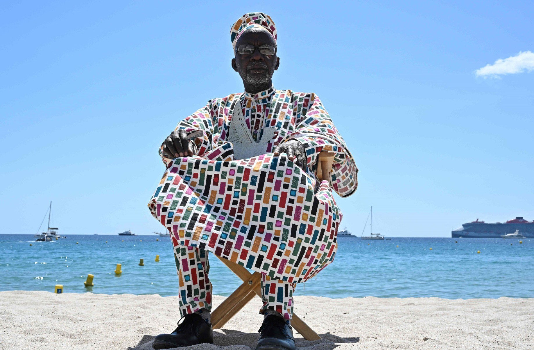 Diretor malinês Souleymane Cissé, vestindo trajes africanos, está sentado em cadeira na praia em Cannes