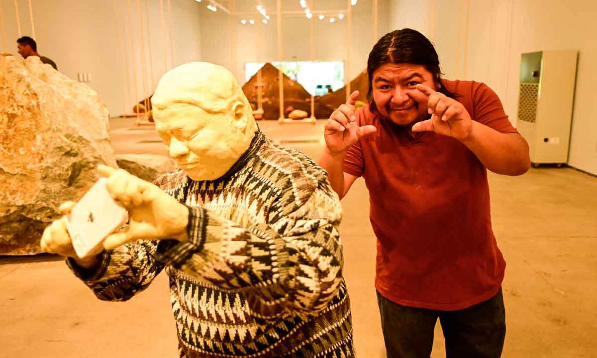 o artista guatemalteco, Edgar Calel na galeria Lago no inhotim, onde está sua exposição. Ele usa camisa vermelha, calça jeans e está diante de uma de suas obras, representada por um homem com uma blusa de lã. O artista faz o gesto como se estivesse fotografando algo.