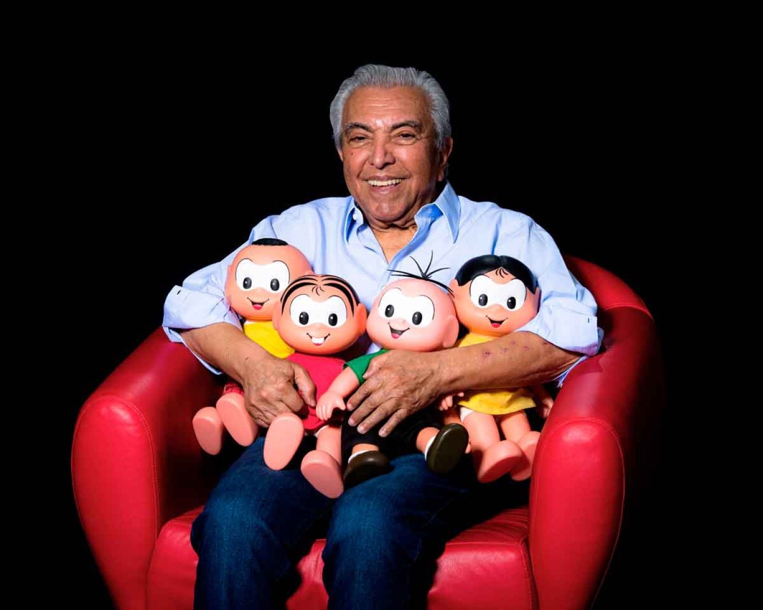 Mauricio de Sousa sorri, sentado na poltrona, abraçado a bonecos de Cascão, Mônica, Cebolinha e Magali