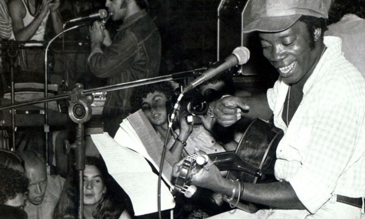 Milton Nascimento est&aacute; no palco e toca viol&atilde;o, sorrindo e sentado em banco, durante show realizado em 1975 em Belo Horizonte