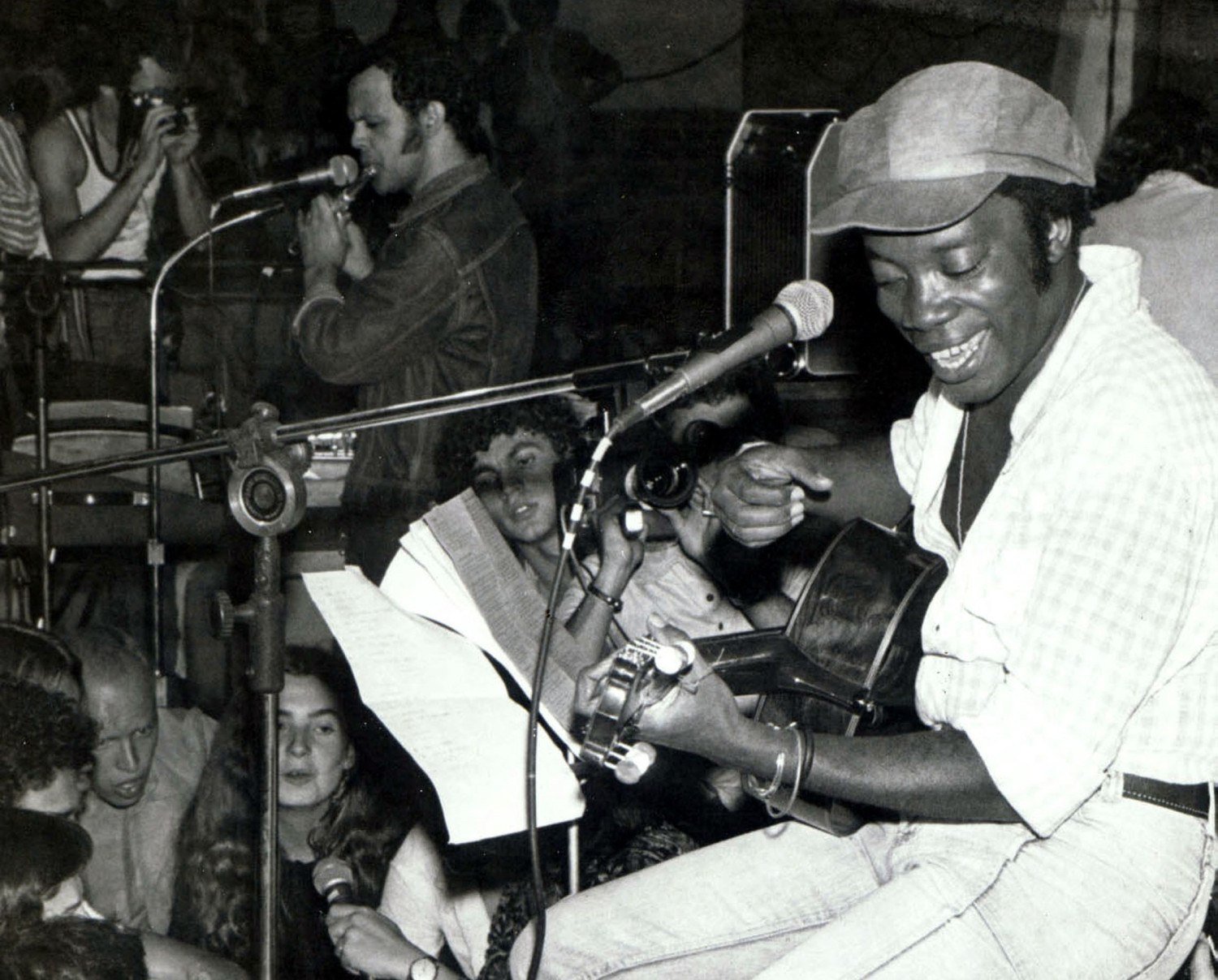 Milton Nascimento está no palco e toca violão, sorrindo e sentado em banco, durante show realizado em 1975 em Belo Horizonte