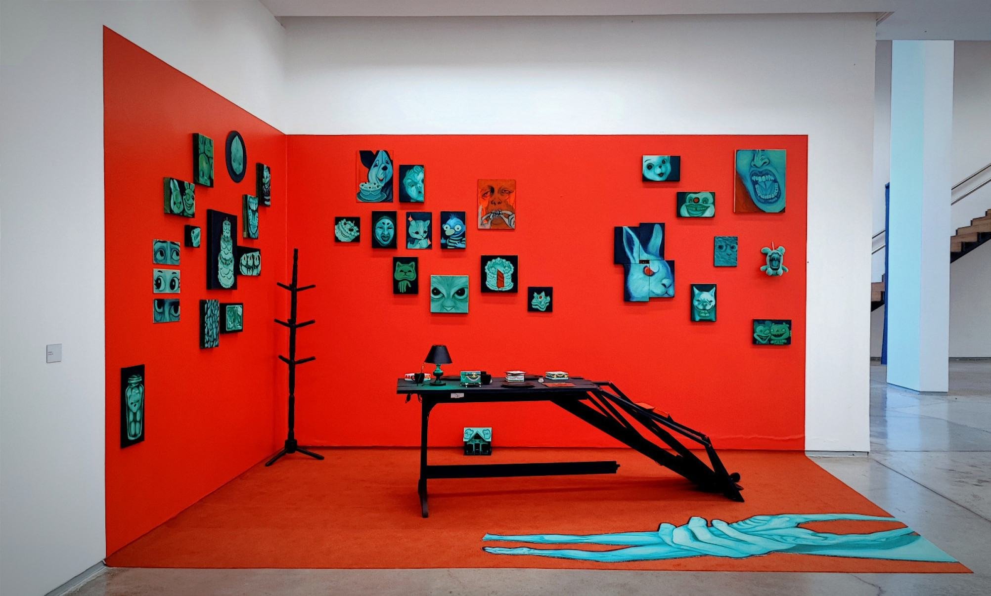  Instala&ccedil;&atilde;o na exposi&ccedil;&atilde;o Estilingue tem c&ocirc;modo na cor vermelha, mesa preta ao centro e quadros com figuras delicadas de gatos e coelhos, al&eacute;m de cenas de pessoas gritando