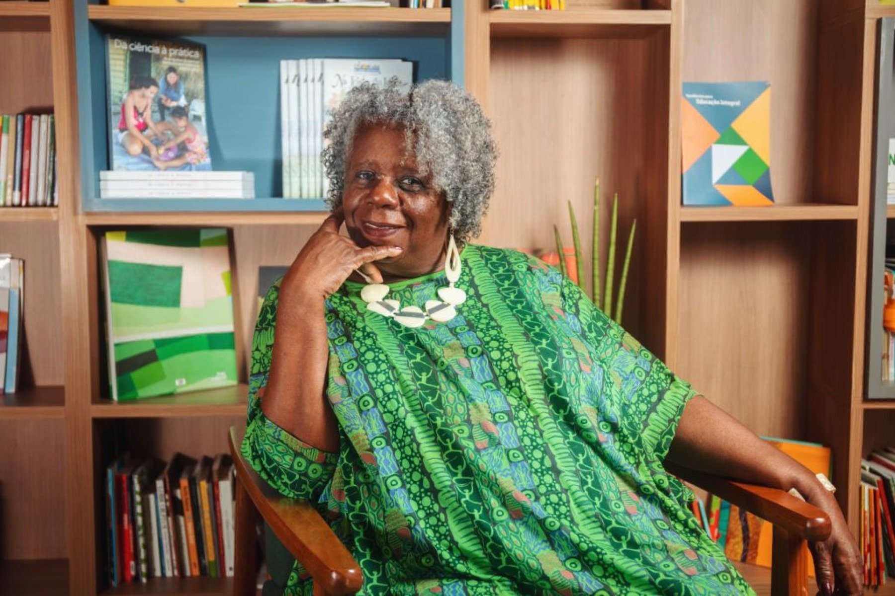 Concei&ccedil;&atilde;o Evaristo usa vestido verde, tem a m&atilde;o no queixo e est&aacute; sentada em frente a uma estante