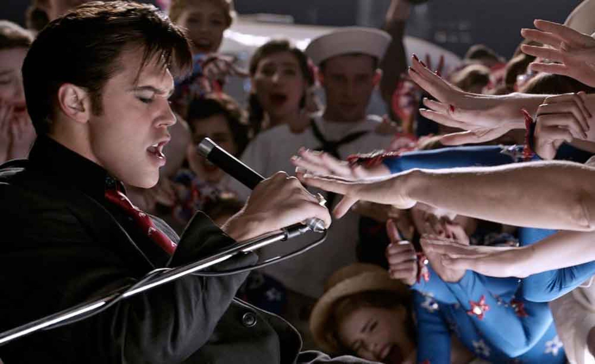 Ator Austin Butler canta ao microfone, no papel de Elvis Presley, diante de m&atilde;os das f&atilde;s