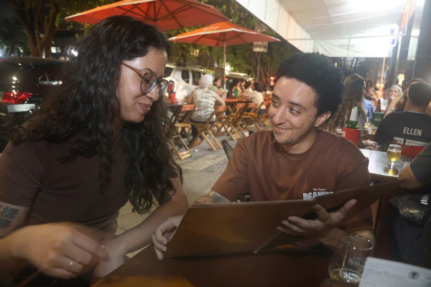 Casal jovem segura card&aacute;pio em bar de Belo Horizonte: eles se olham e, ao fundo, v&ecirc;-se outros clientes sentados em mesas na cal&ccedil;ada