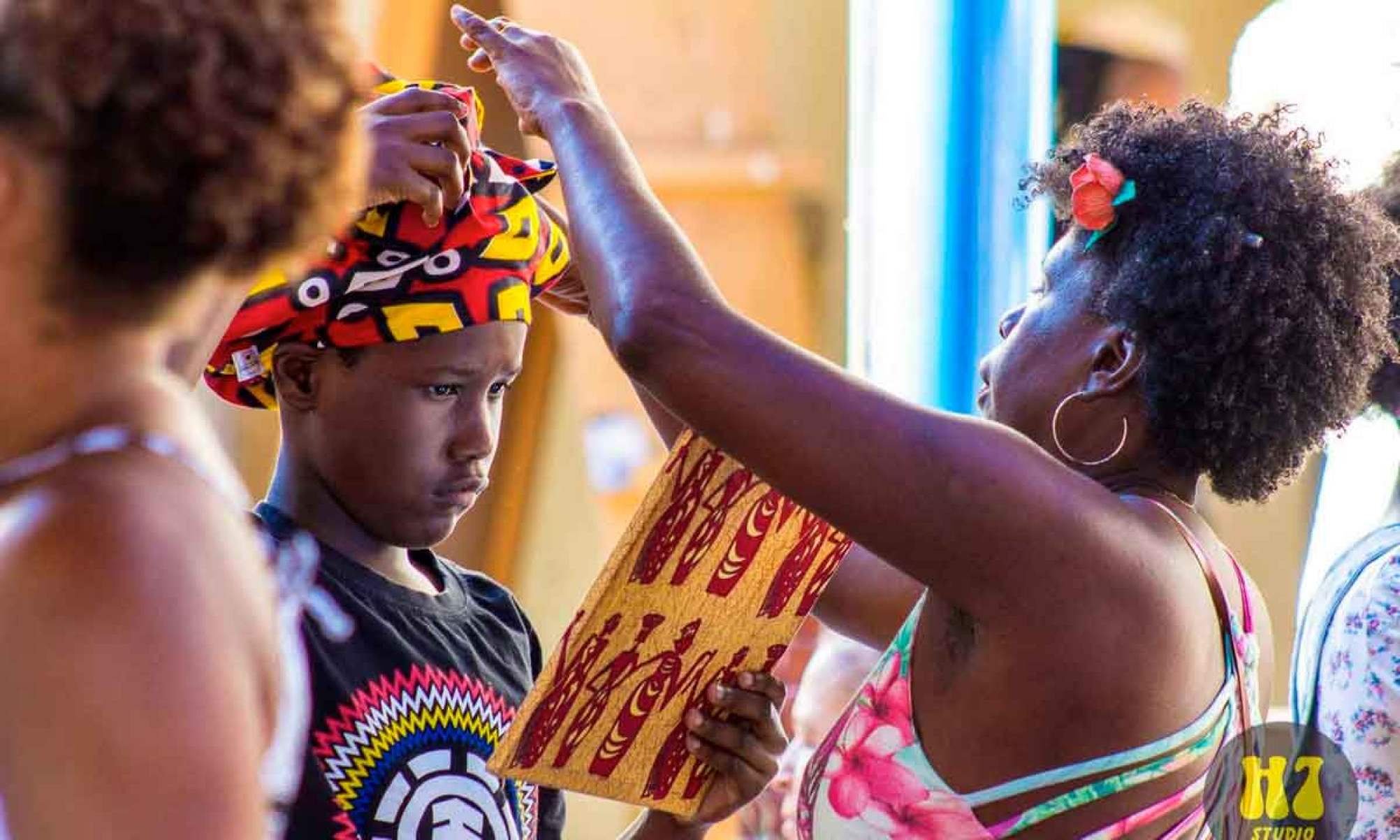 Mulher com flor no cabelo ajeita turbante colorido de crian&ccedil;a, que l&ecirc; papel 