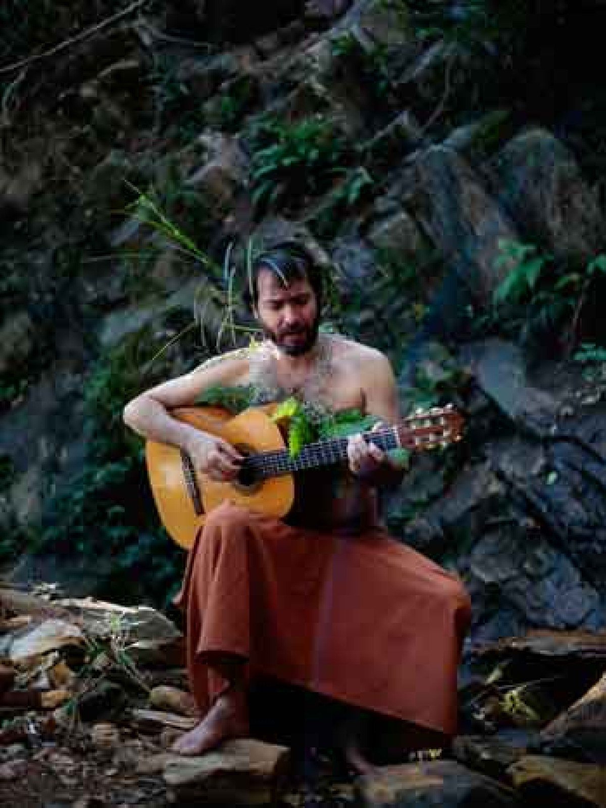 Sem camisa e com saia marrom, o m&uacute;sico Leandro C&eacute;sar toca viol&atilde;o sentado em pedra, com pared&atilde;o rochoso atr&aacute;s de si