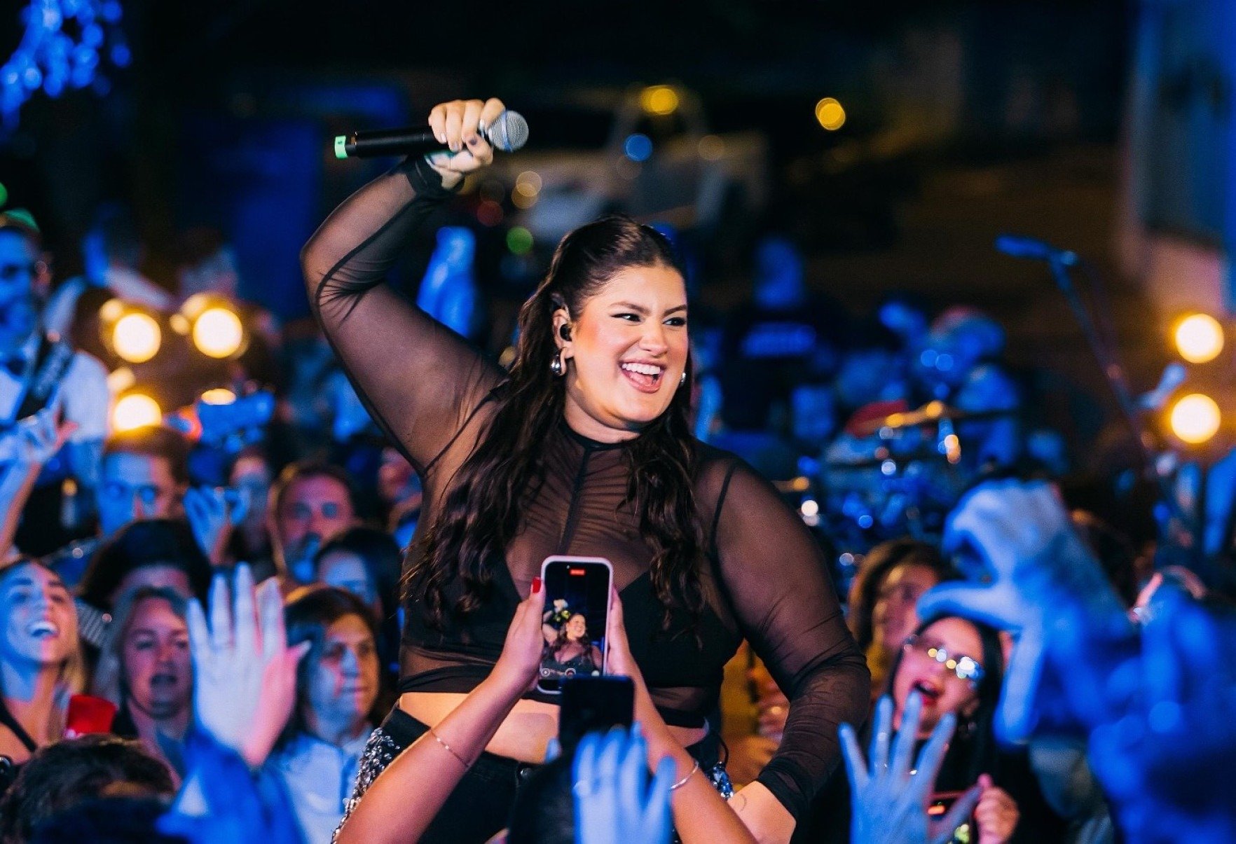 Cantora Bruna Lipiani sorri, com o microfone na mão, cercada de mãos erguendo celulares para fotografá-la