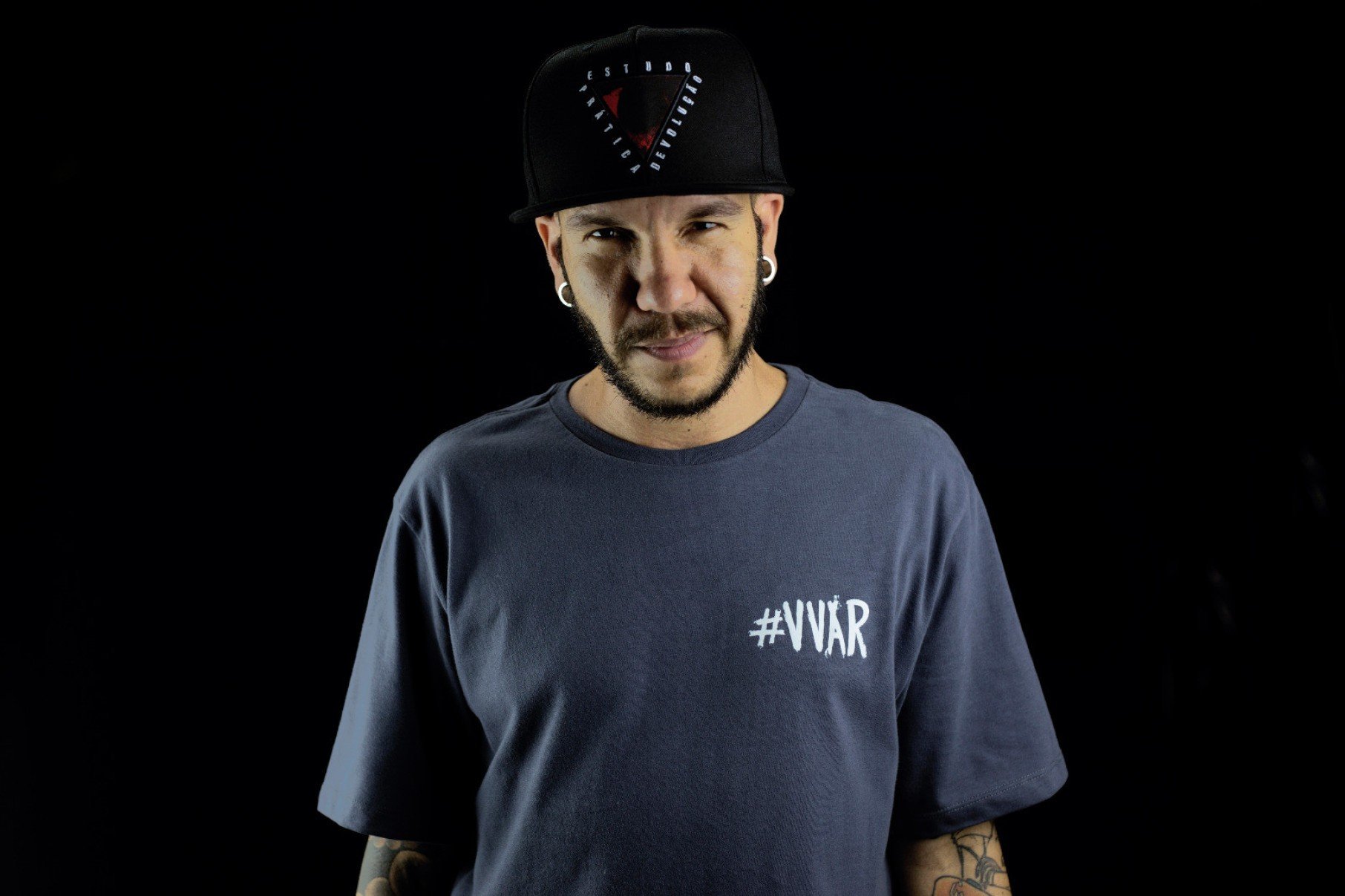 Rapper Monge usa blusa azul-marinho e olha para a câmera 