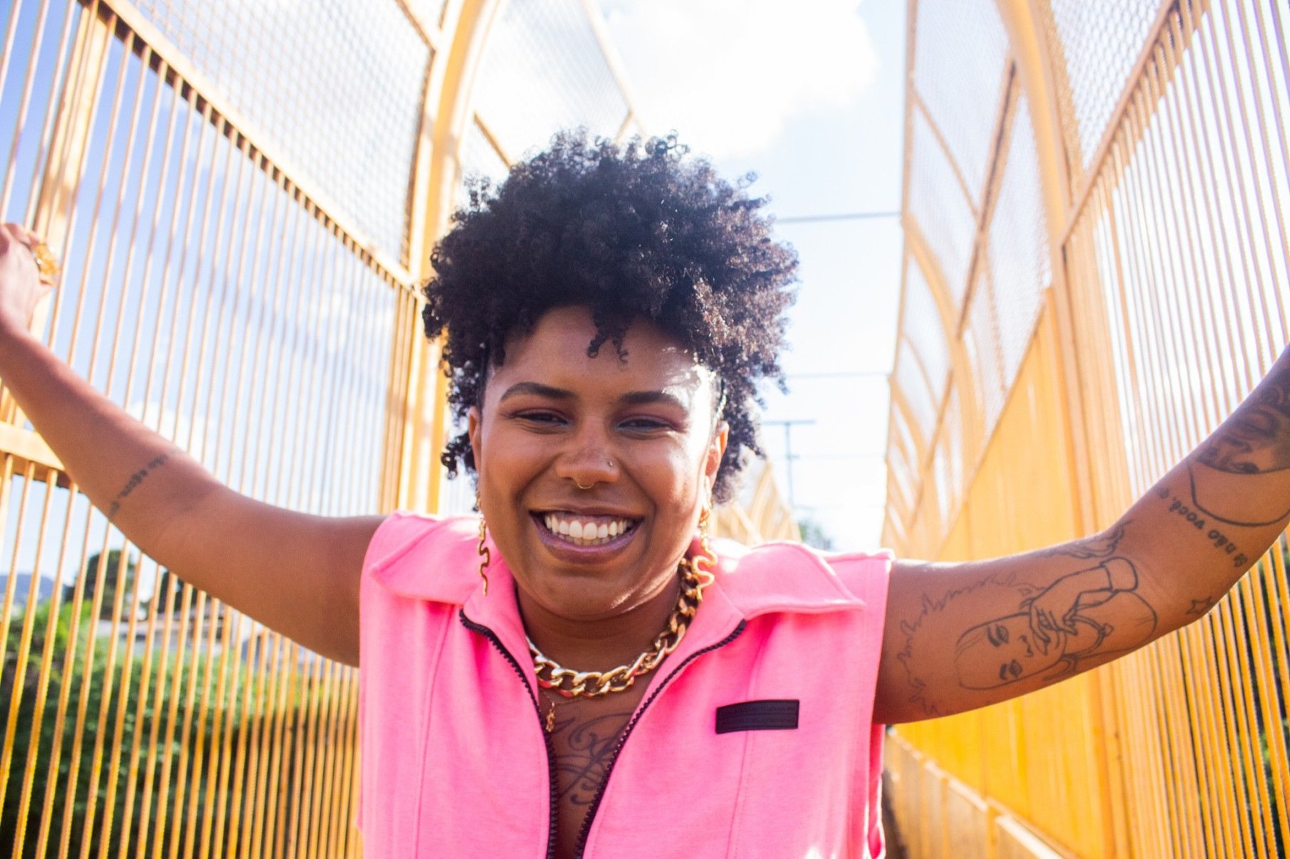 Cantora e compositora Ohana sorri, usa blusa rosa e olha para a câmera, com os braços abertos