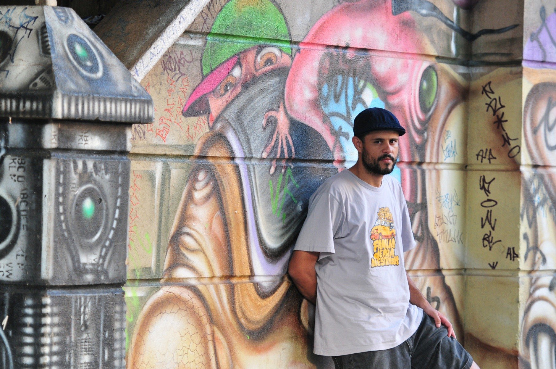 Rapper PDR está encostado em muro pichado com grafite colorido e olha para a câmera