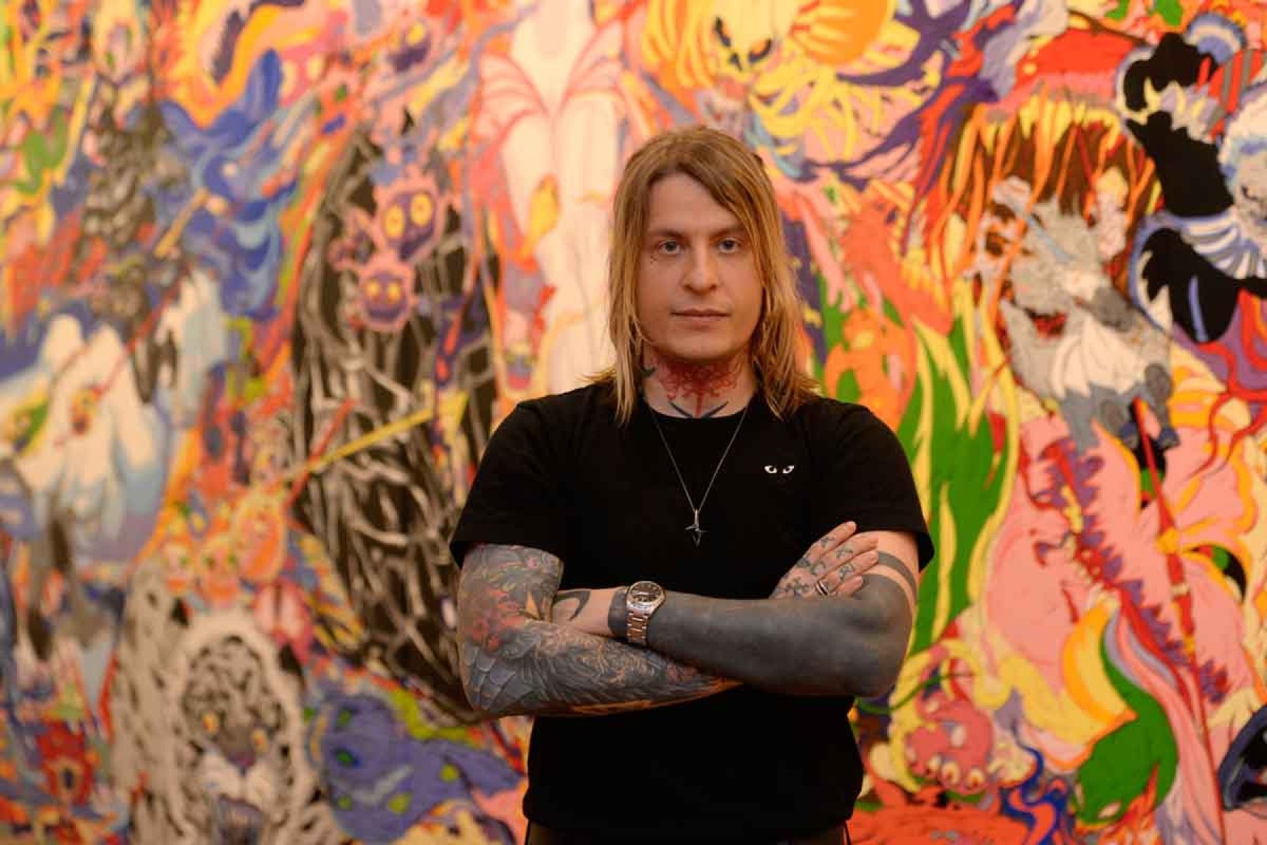 Curador Antonio Curti olha para a câmera. Tem cabelos louros longos e está de braço cruzado, usando camiseta preta