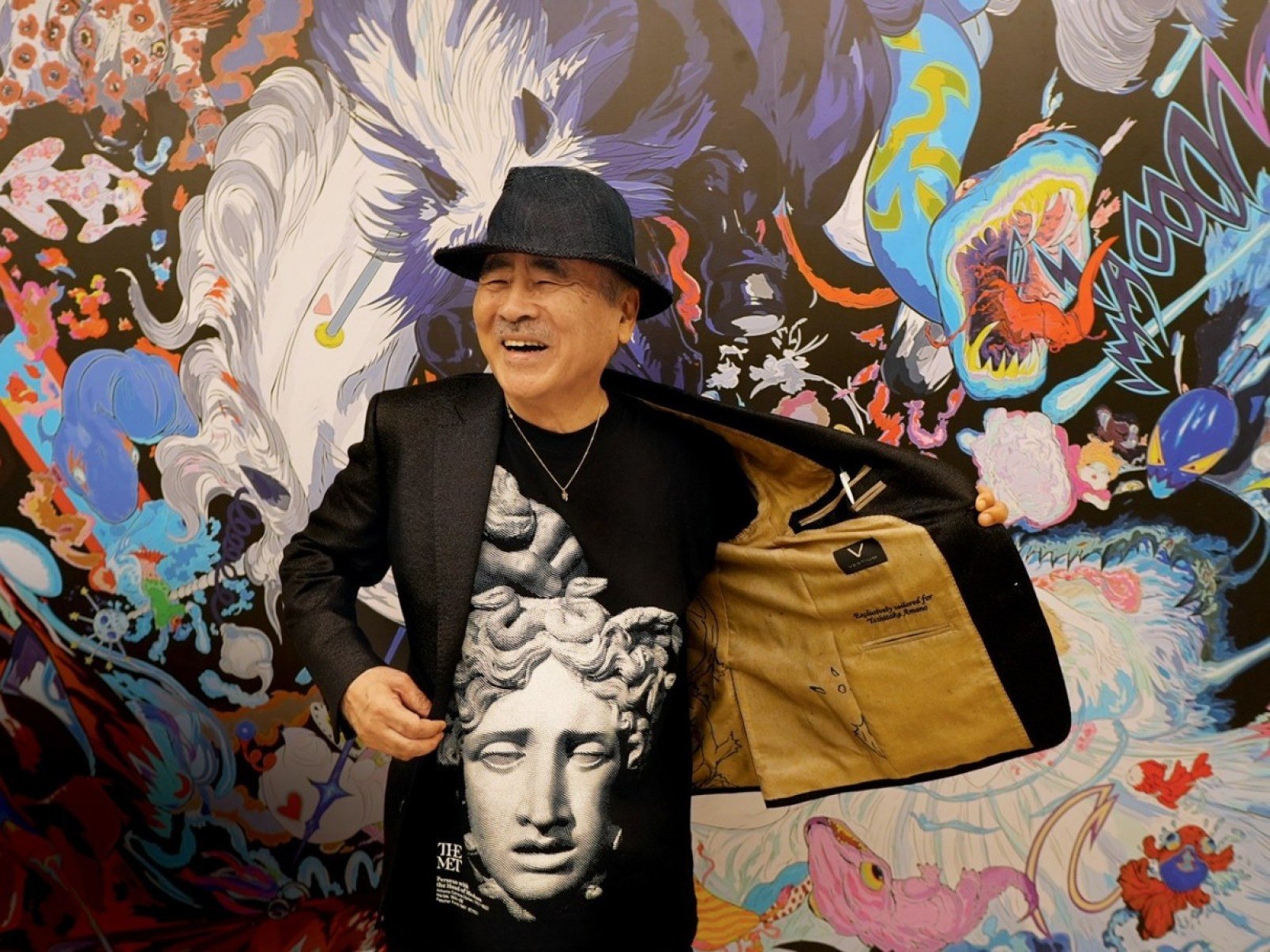  Artista japonês Yoshitaka Amano sorri, abre o paletó preto que está vestindo e exibe camiseta estampada com figura feminina de estátua da Antiguidade. Ao fundo, há parede com painel multicolorido 