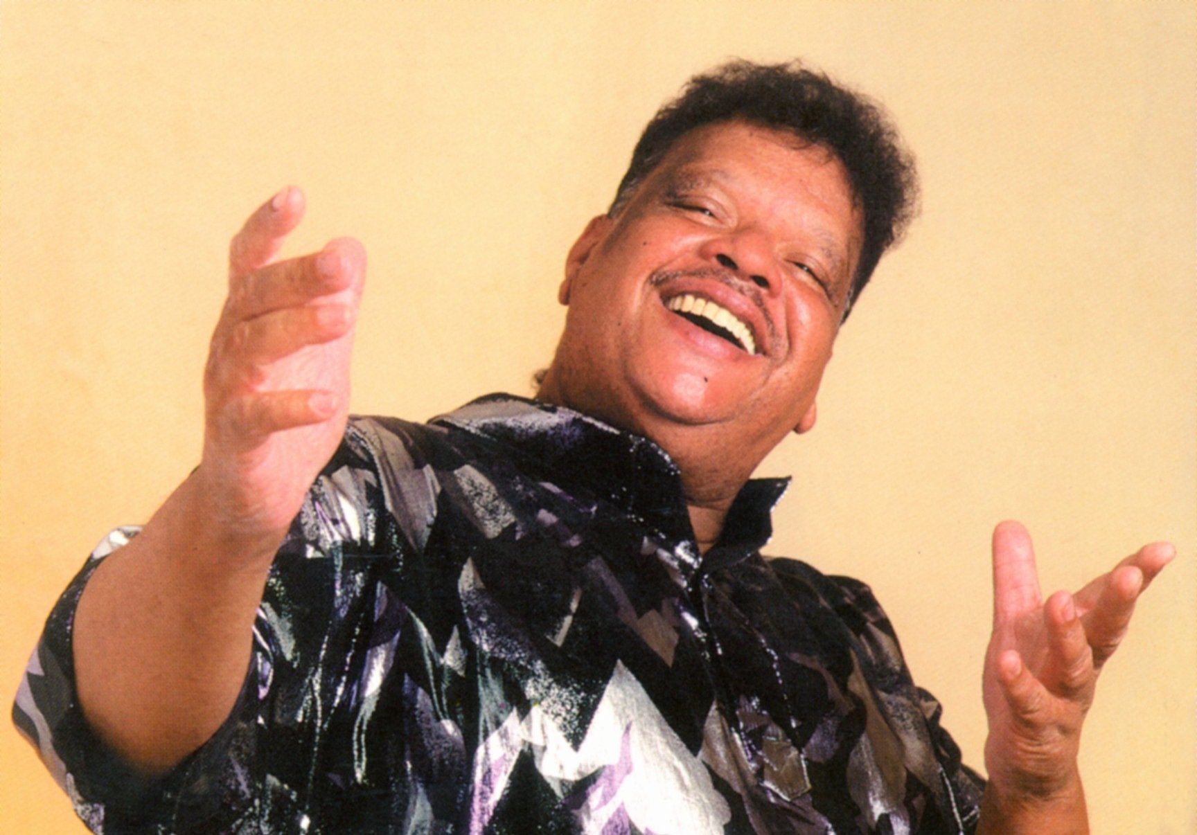 Tim Maia olha para a câmera sorrindo com os braços abertos e as mãos em frente ao corpo