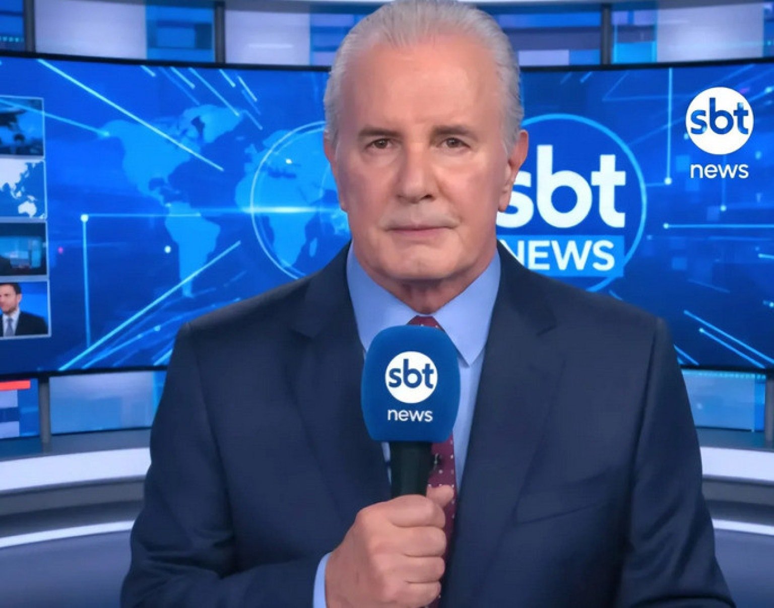 Celso Freitas segura o microfone e atrás dele telão azul exibe a logomarca do SBT News