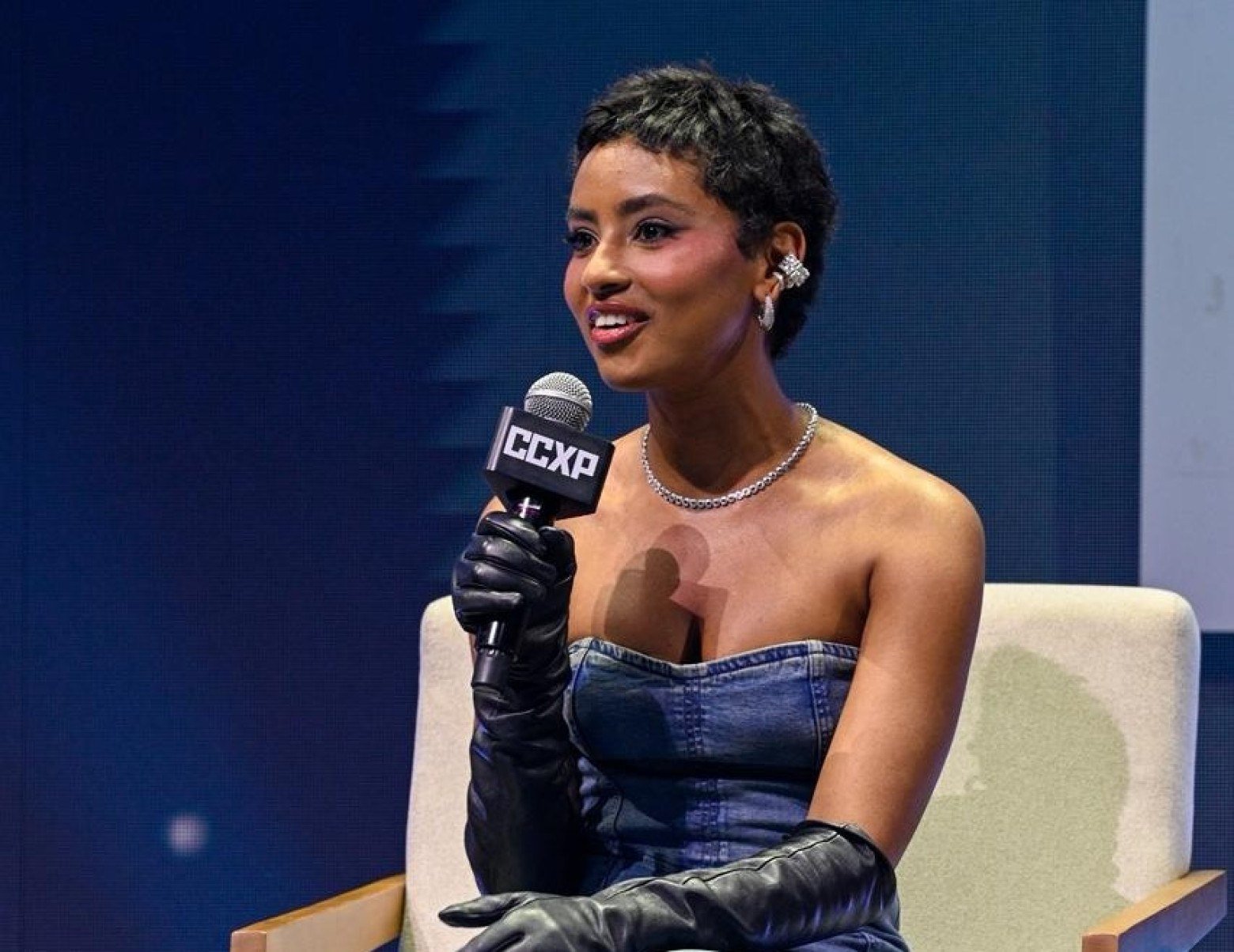 Gabz está sentada, de vestido tomara que caia e luvas. Ela fala ao microfone durante painel na CCXP