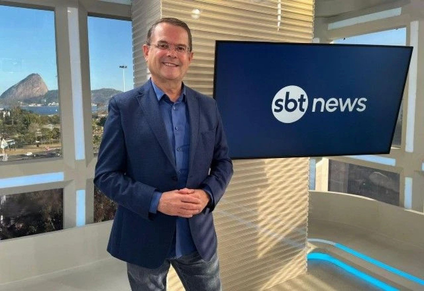 O jornalista Sidney Rezende sorri ao lado da tela onde se vê a logomarca do canal SBT News. Através da janela, ao fundo, vê-se o Pão de Açúcar em dia ensolorado