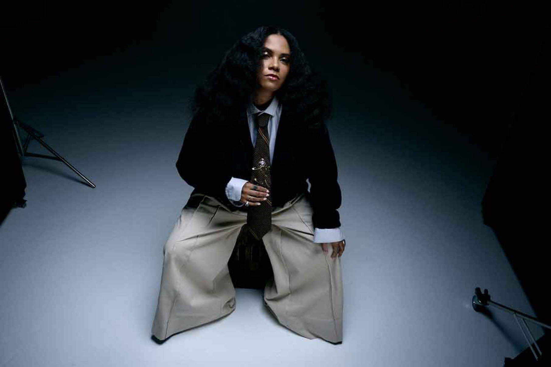 DJ Kingdom é fotografada do alto, sentada em um banco com jaqueta de mangas compridas e calças muito largas