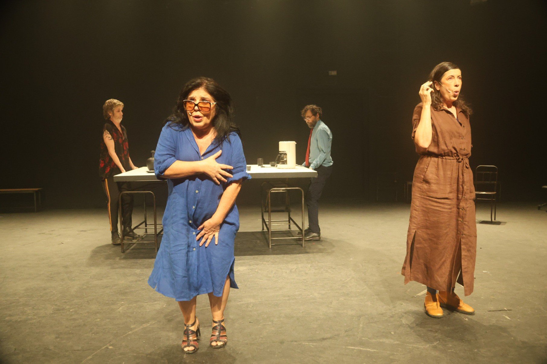 Atriz de &oacute;culos e vestido azul e atriz de vestido marrom est&atilde;o &agrave; frente, dois atores est&atilde;o em mesa, ao fundo, durante ensaio da pe&ccedil;a Um ensaio sobre a cegueira, no Galp&atilde;o Cine Horto, em BH