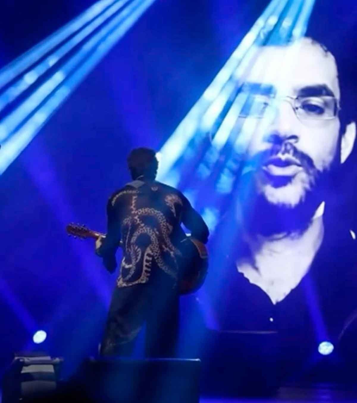 Cantor Paulo Ricardo est&aacute; de costas, tocando guitarra, de frente para imenso tel&atilde;o com a imagem de Renato Russo 