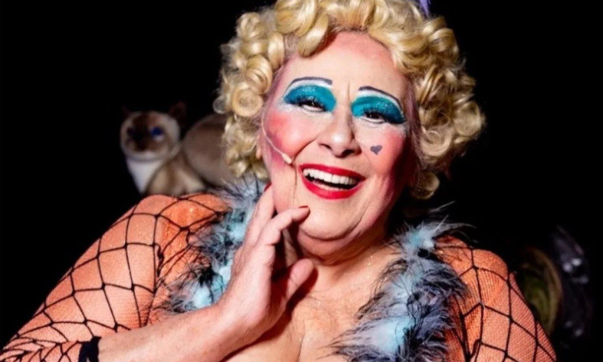 Teuda Bra sorri, com rosto fortemente maquiado e peruca loura, caracterizada como personagem do Cirque du Soleil