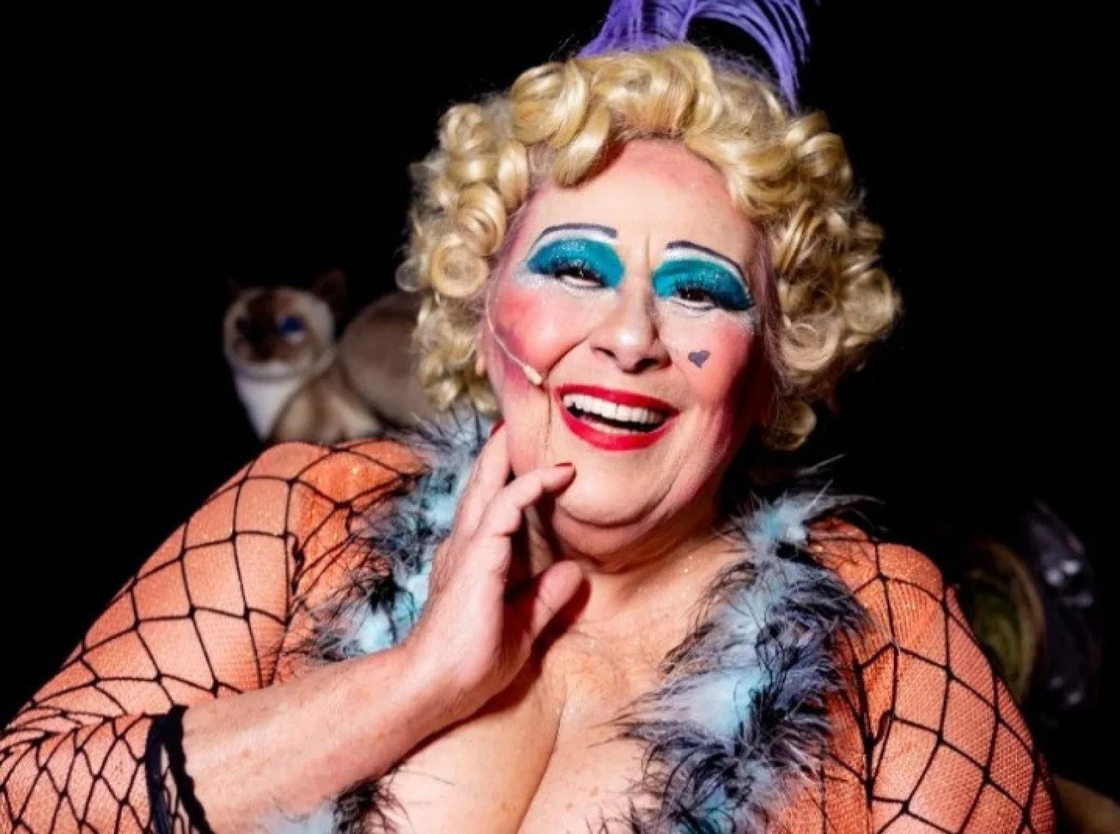 Teuda Bra sorri, com rosto fortemente maquiado e peruca loura, caracterizada como personagem do Cirque du Soleil