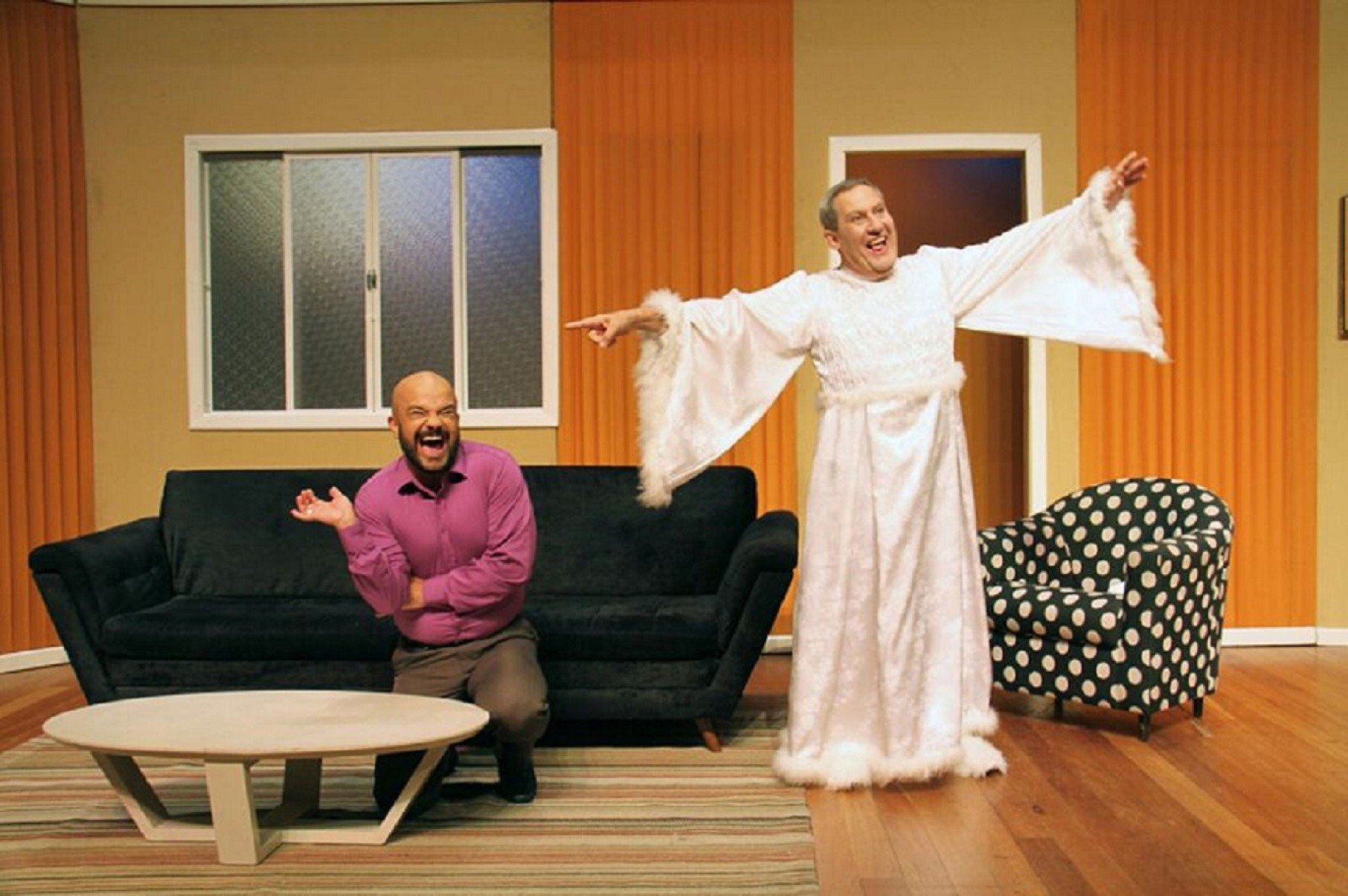 Ator Mauricio Cangu&ccedil;u est&aacute; sentado no sof&aacute; e ator Ilvio Amaral, vestido de fantasma, est&aacute; ao lado dele, de p&eacute;, com os bra&ccedil;os abertos, em cena da pe&ccedil;a Acredite, um esp&iacute;rito baixou em mim