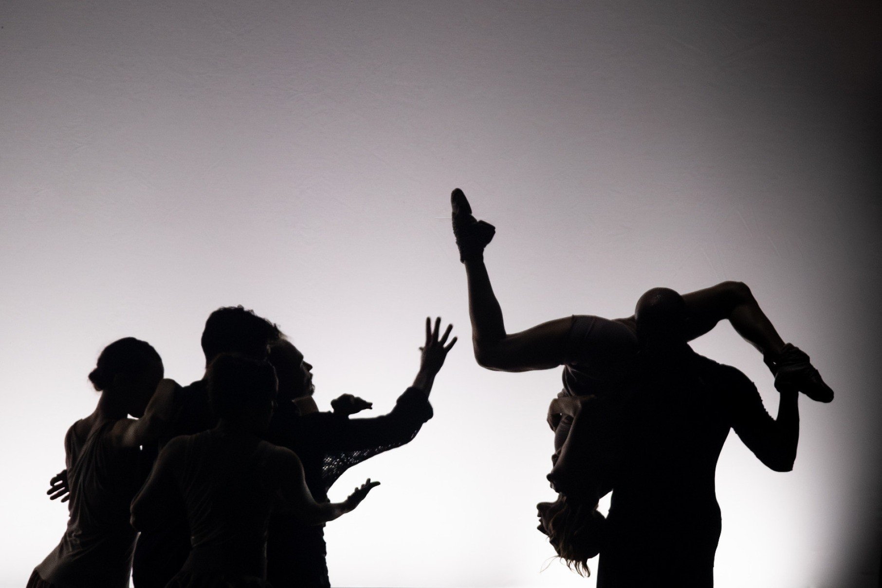 Bailarinos da companhia Mimulus est&atilde;o em p&eacute;, contra a luz. Um deles leva nos ombros bailarina de cabe&ccedil;a para baixo com o tornozelo erguido para o teto