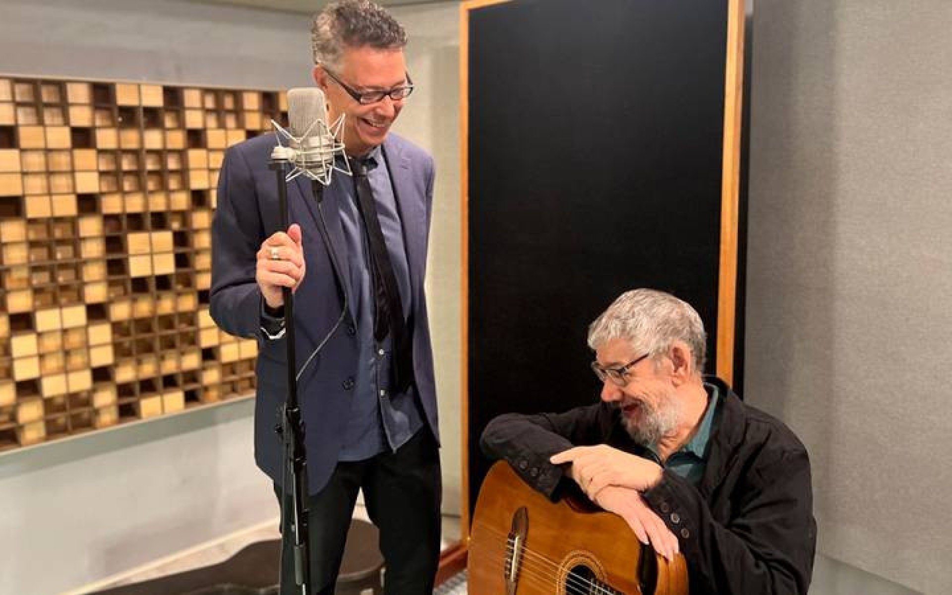 Augusto Martins est&aacute; em p&eacute; segurando o microfone ao lado de H&eacute;lio Delmiro, sentado, segurando o viol&atilde;o