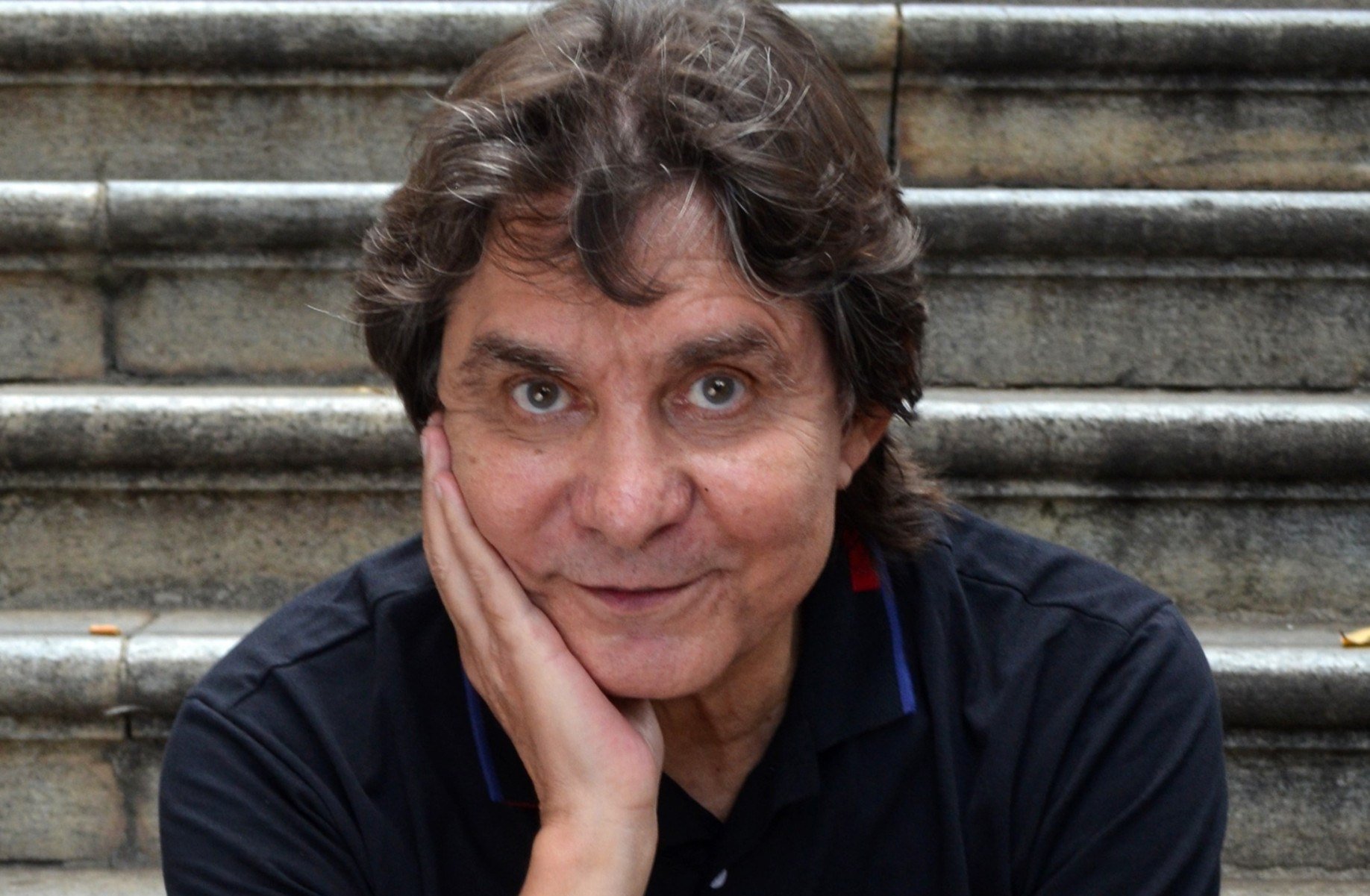 L&ocirc; Borges usa camiseta preta, olha para a c&acirc;mera e tem a m&atilde;o direita no rosto. Atr&aacute;s dele se veem degraus de uma escada ao ar livre