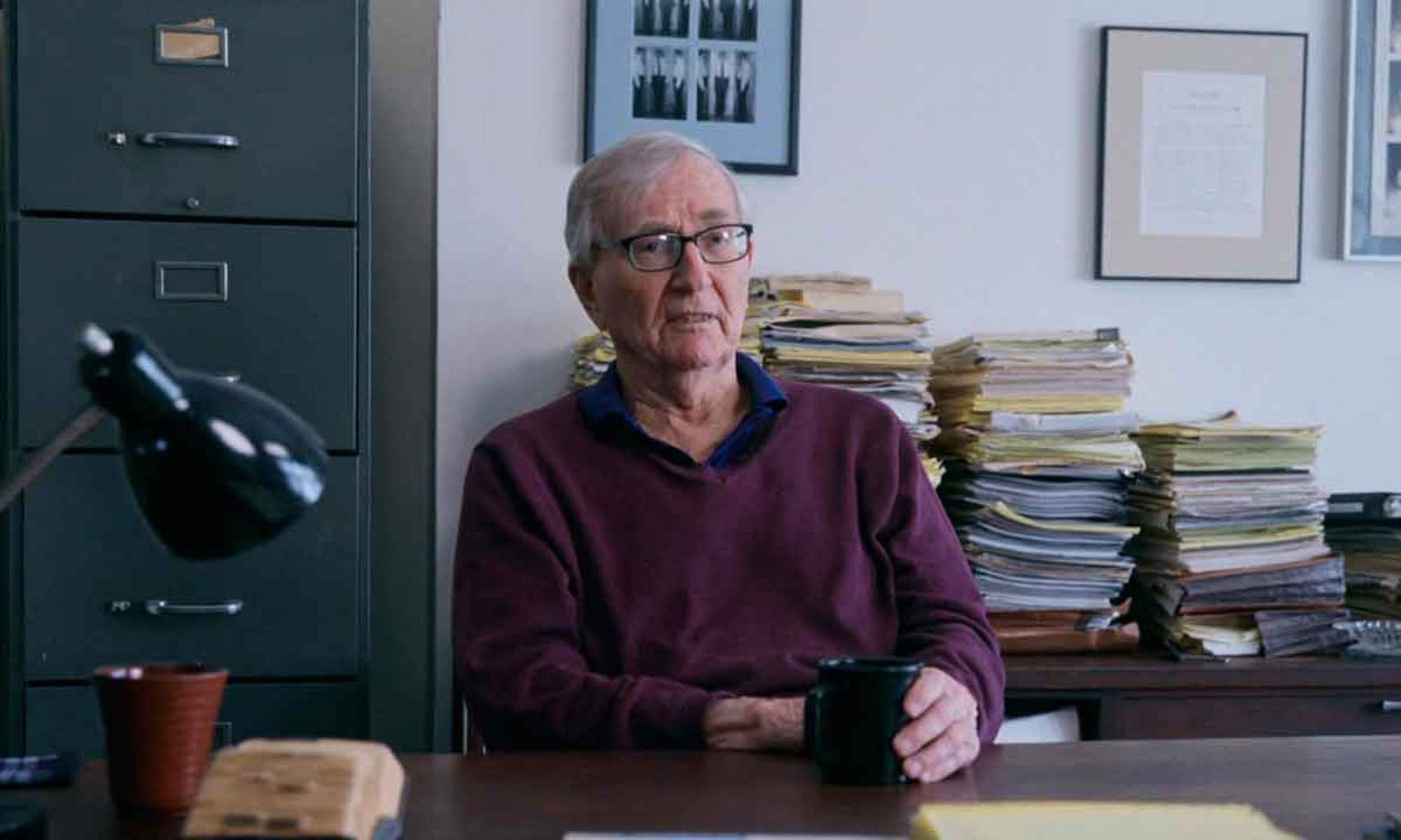 Fot&oacute;grafo Seymour Hersh est&aacute; sentado em mesa de trabalho, tendo atr&aacute;s de si pilhas de papel, em cena do filme "Seymour Hersh: em busca da verdade"