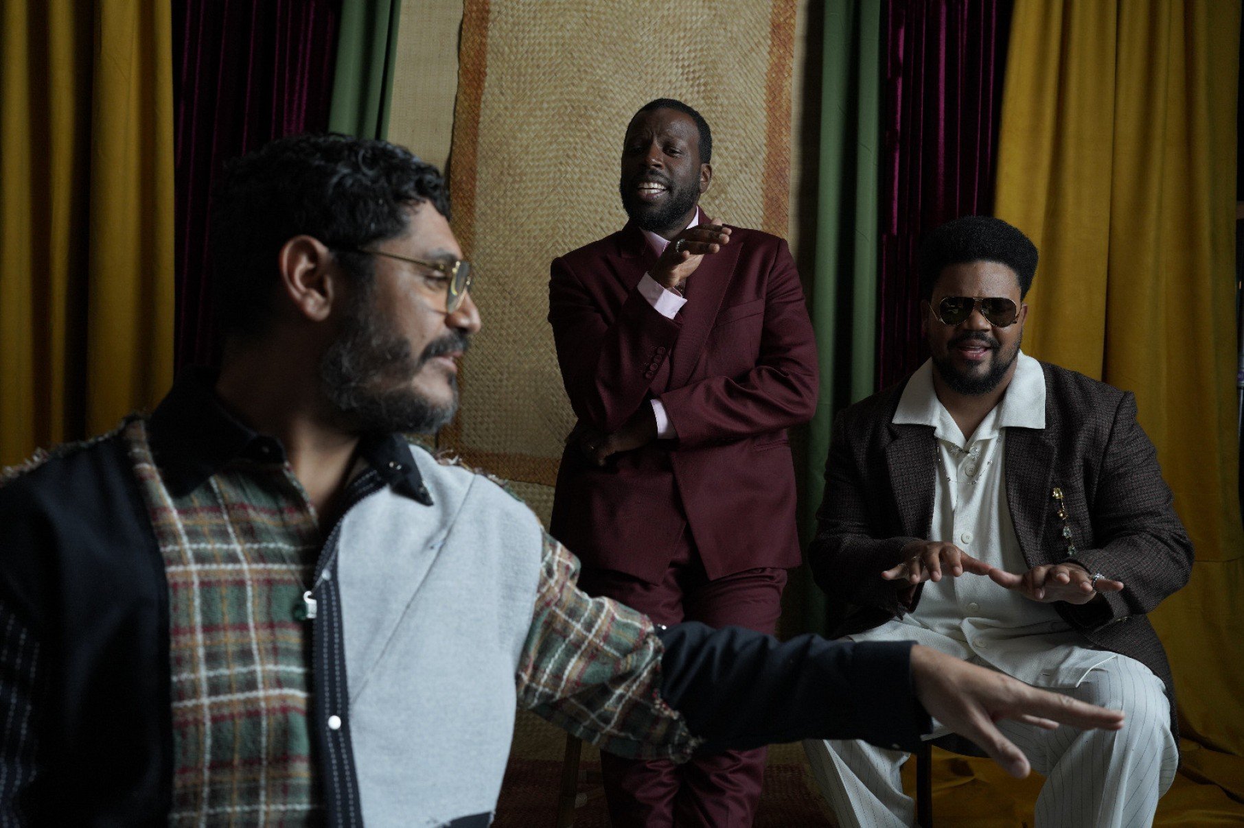   M&uacute;sico Criolo est&aacute; de lado com a m&atilde;o estendida, em primeiro plano. Atr&aacute;s dele, de terno vinho Dino D'Santiago tem a m&atilde;o no queixo. &Agrave; direita, Amaro Freitas faz gesto com as m&atilde;os fingindo tocar piano