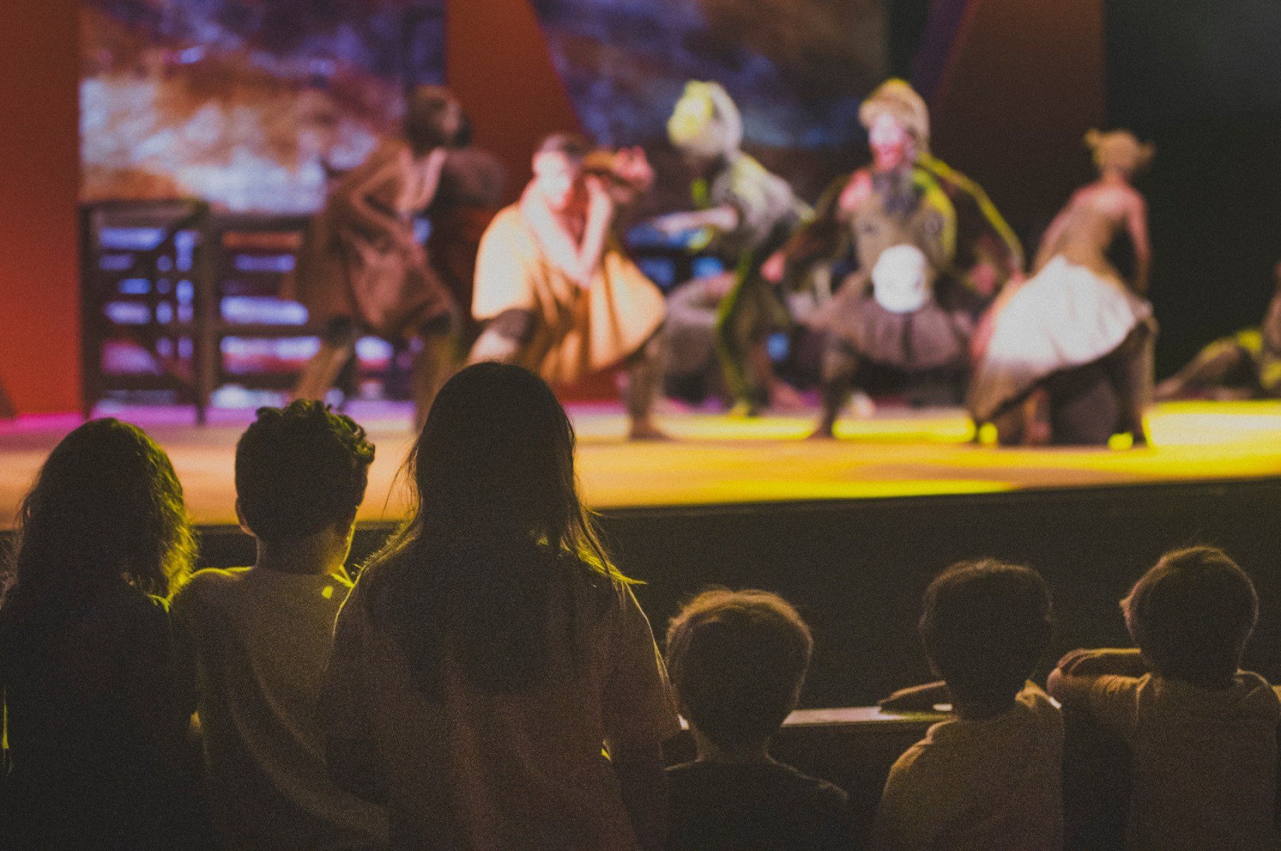 Crian&ccedil;as assistem no escuro &agrave; pe&ccedil;a Os saltimbancos. Elas est&atilde;o em primeiro plano, de costas para a c&acirc;mera. Ao fundo, veem-se artistas no palco colorido e iluminado