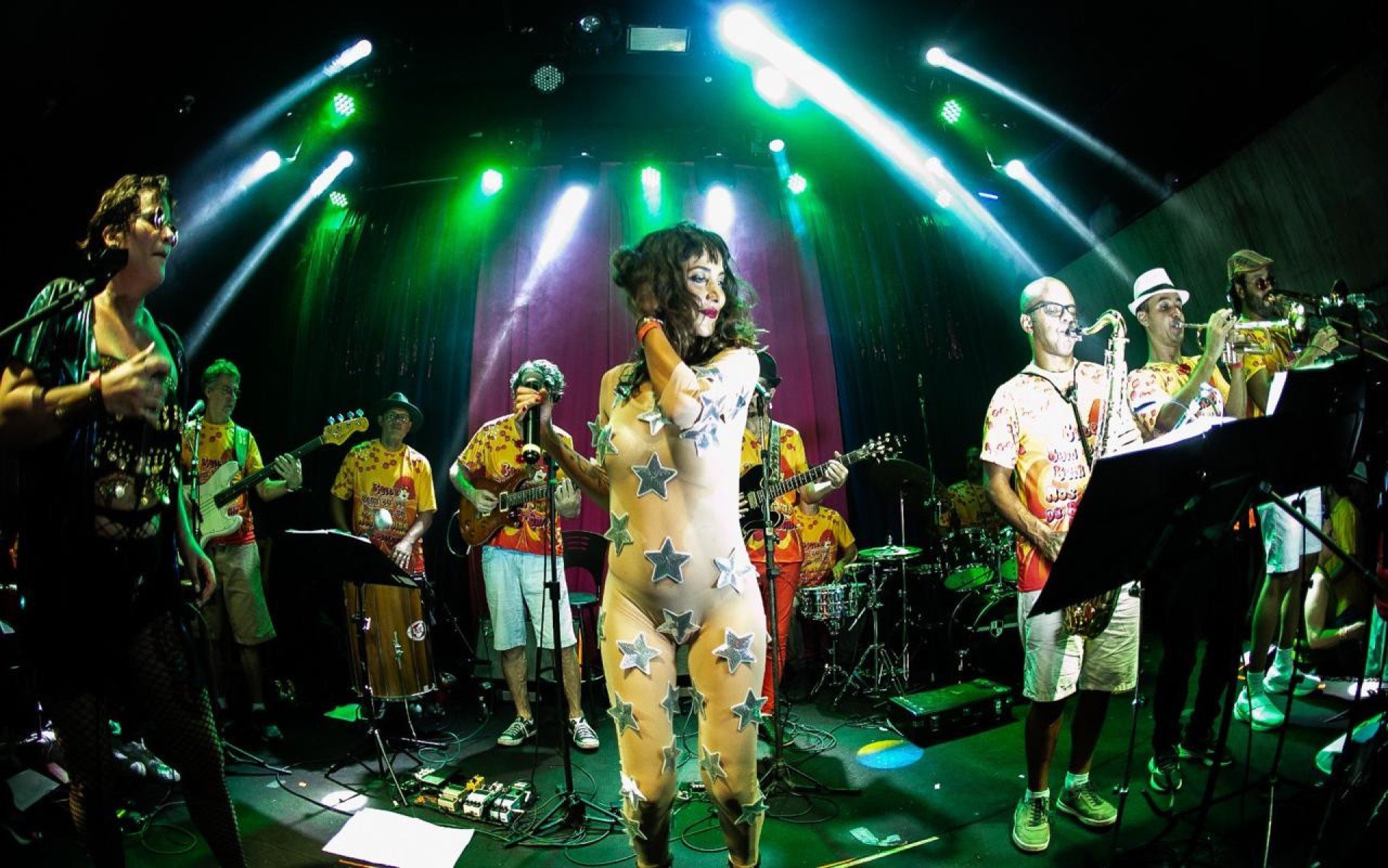 Cantora vestida com macac&atilde;o com estrelas prateadas segura o microfone, no centro do palco, cercada por oito m&uacute;sicos que tocam guitarra e sopros