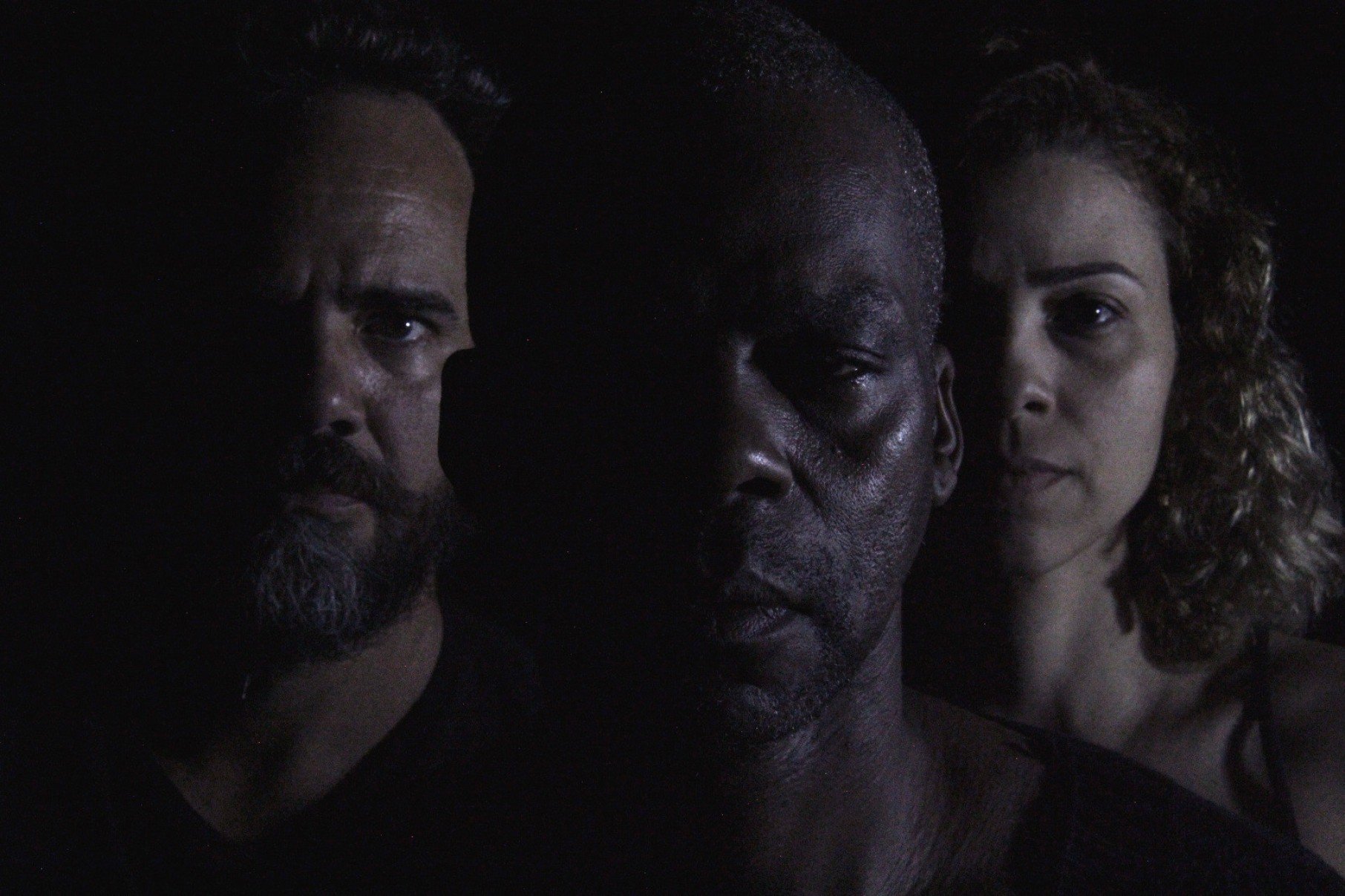 Close dos rostos de tr&ecirc;s personagens da pe&ccedil;a Otelo. Ator negro que interpreta Otelo est&aacute; no centro, ladeado por um ator de barba, &agrave; esquerda, e atriz &agrave; direita