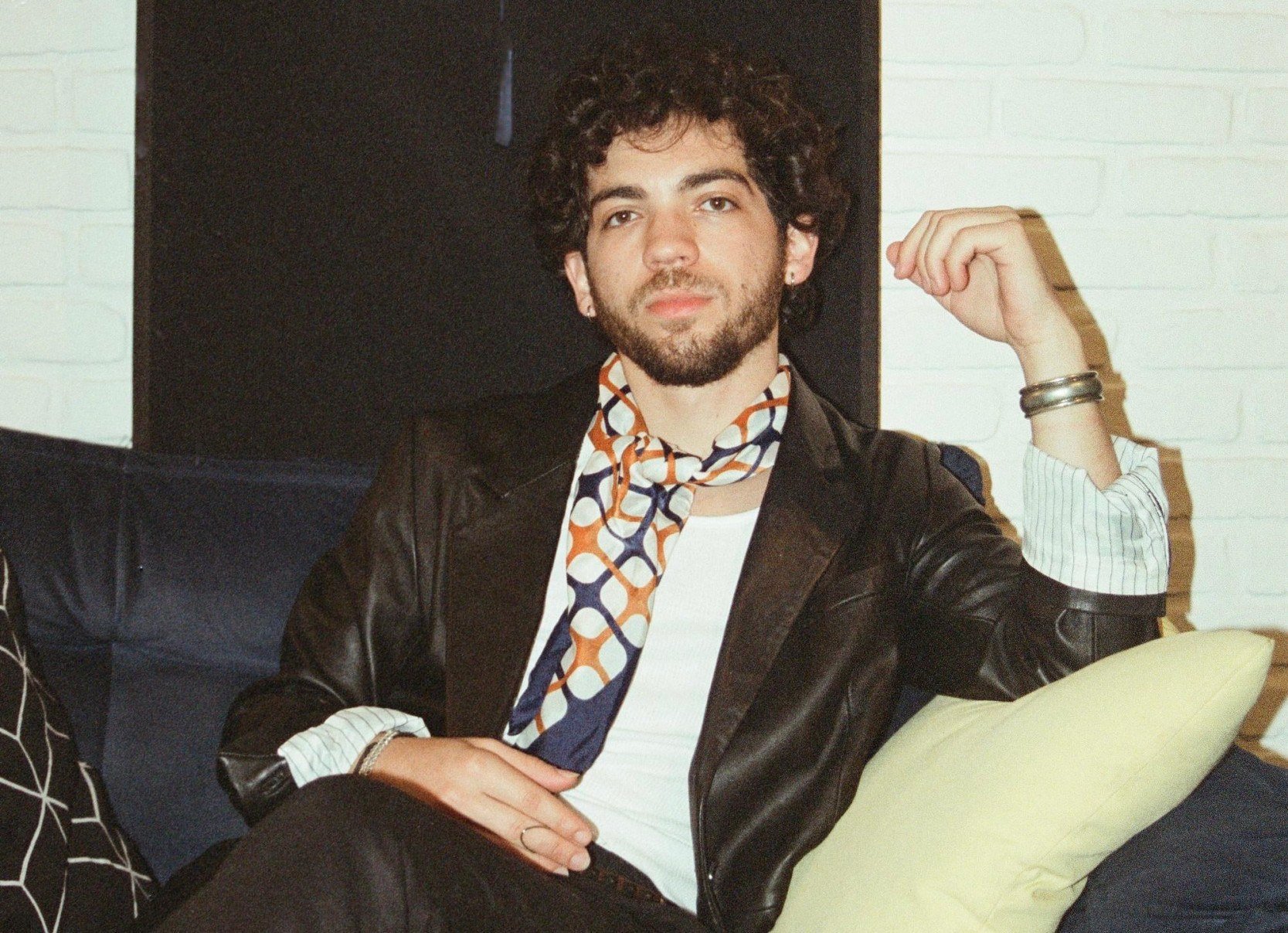 Compositor Davi Le&atilde;o olha para a c&acirc;mera. Ele est&aacute; sentado em sof&aacute; escuro, veste casaco escuro, camisa branca e tem len&ccedil;o estampado no pesco&ccedil;o