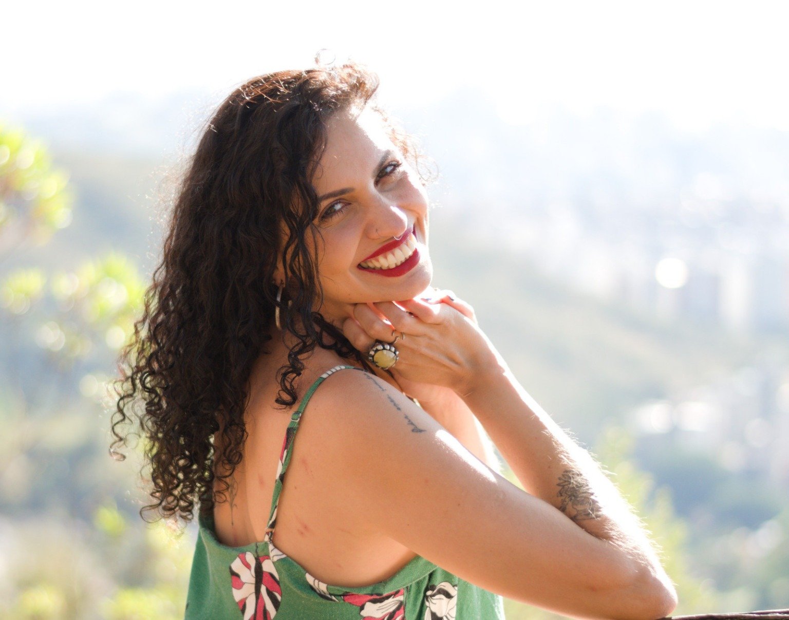 Compositora My Let&iacute;cia est&aacute; sentada em banco de madeira, olha para tr&aacute;s com a m&atilde;o na queixo. Atr&aacute;s dela v&ecirc;-se a paisagem ensolarada de BH