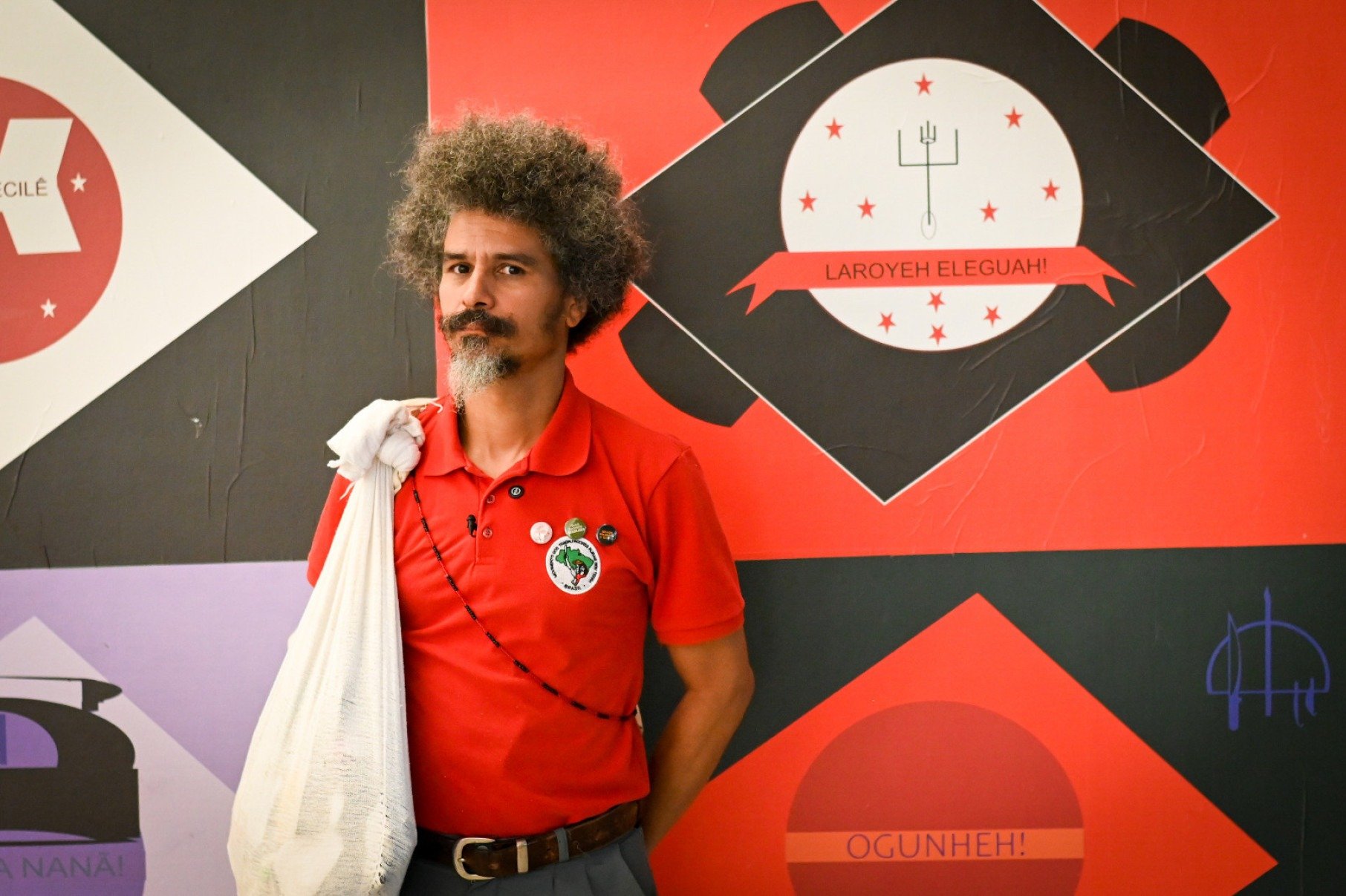 O artista Paulo Nazareth olha para a c&acirc;mera. Ele usa blusa vermelha e est&aacute; &agrave; frente de pintura em tons vermelhos na Galeria Pra&ccedil;a, em Inhotim, Minas Gerais