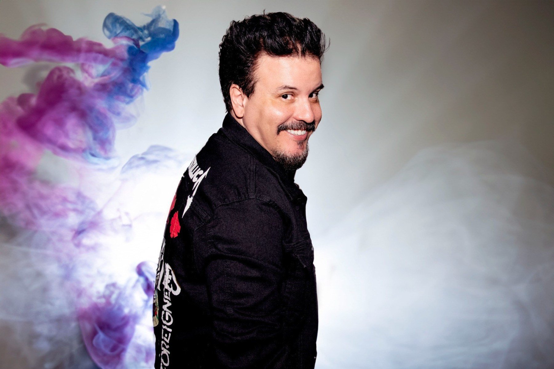 Luciano Nassyn, que foi cantor do grupo Trem da Alegria, veste camisa preta, est&aacute; de costas, de lado, e sorri para a c&acirc;mera