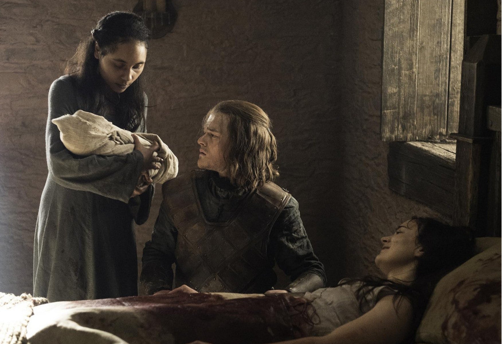 Mulher est&aacute; de p&eacute; e segura beb&ecirc; observado pelo ator Robert Aramayo, ajoelhado. Na frente dele, deitada, est&aacute; a m&atilde;e da crian&ccedil;a, que acabou de dar &agrave; luz