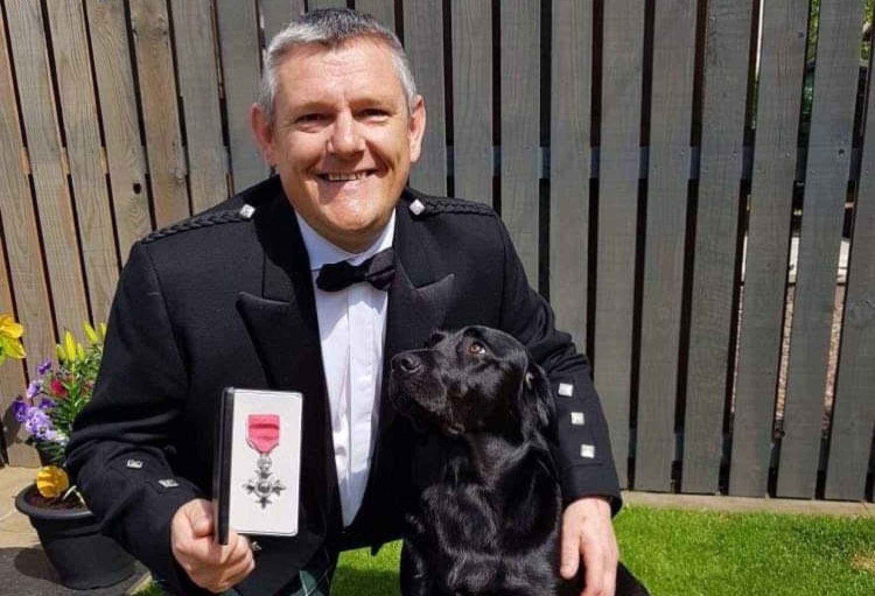 John Davidson, militante da campanha de esclarecimento sobre a S&iacute;ndrome de Tourette, com a medalha que recebeu da rainha Elizabeth II, em 2019. Ele sorri para a c&acirc;mera e abra&ccedil;a um cachorro preto