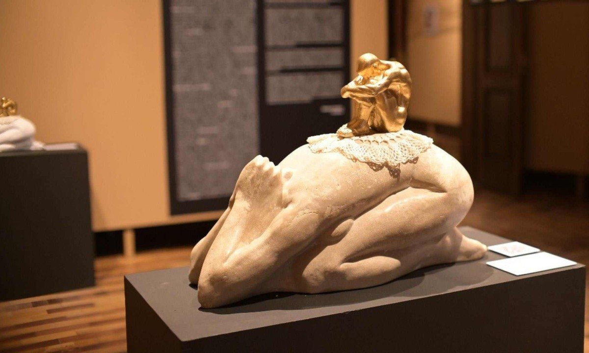 Escultura branca criada por Marlene Barros mostra mulher de joelhos, com o rosto entre os bra&ccedil;os e em posi&ccedil;&atilde;o humilhante. Em cima das costas dela h&aacute; pequena escultura dourada de figura masculina sobre paninho de croch&ecirc;